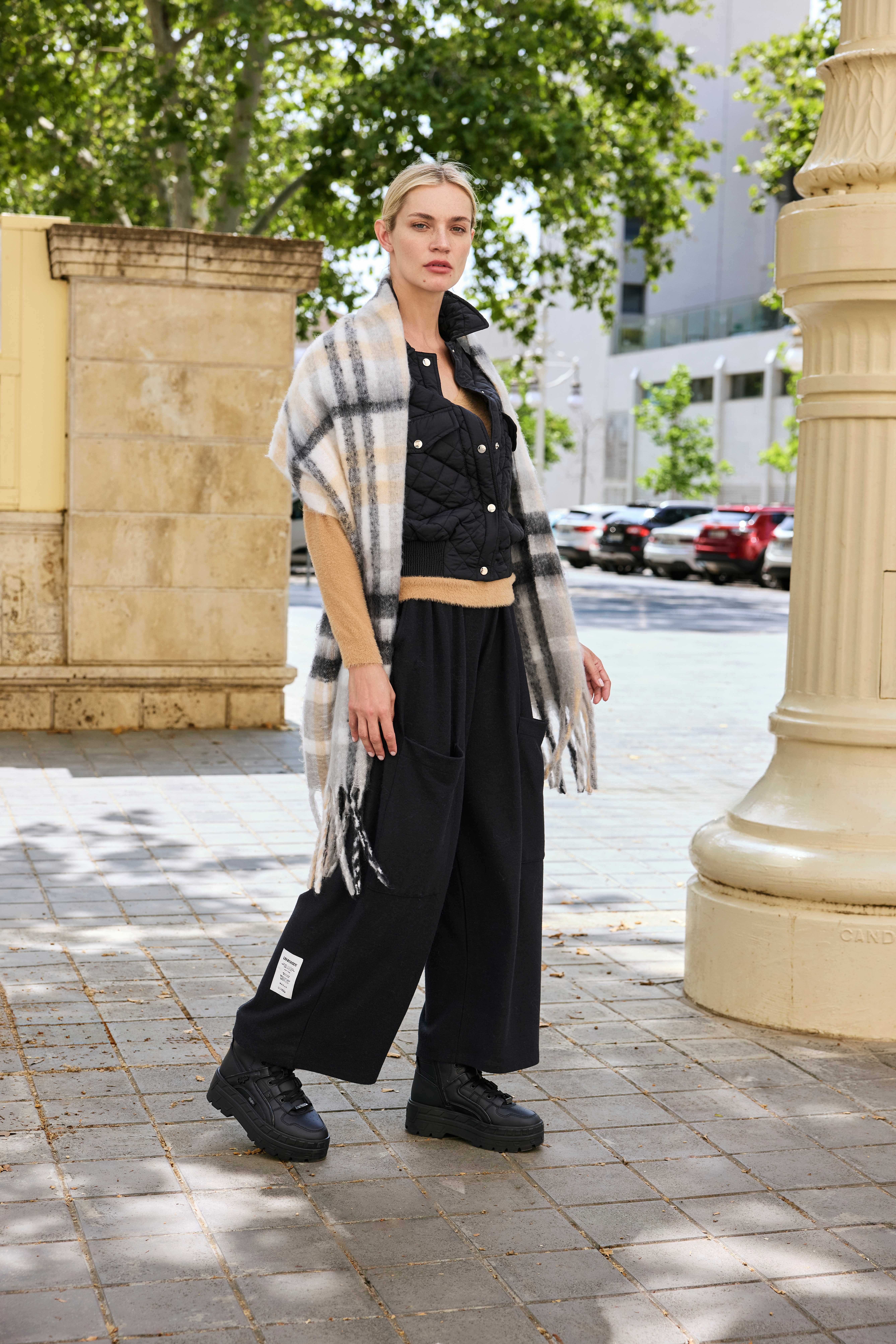 Blonde Frau mit schwarzem Knopf-Top, langer schwarzer Hose und passenden Boots, in urbaner Umgebung