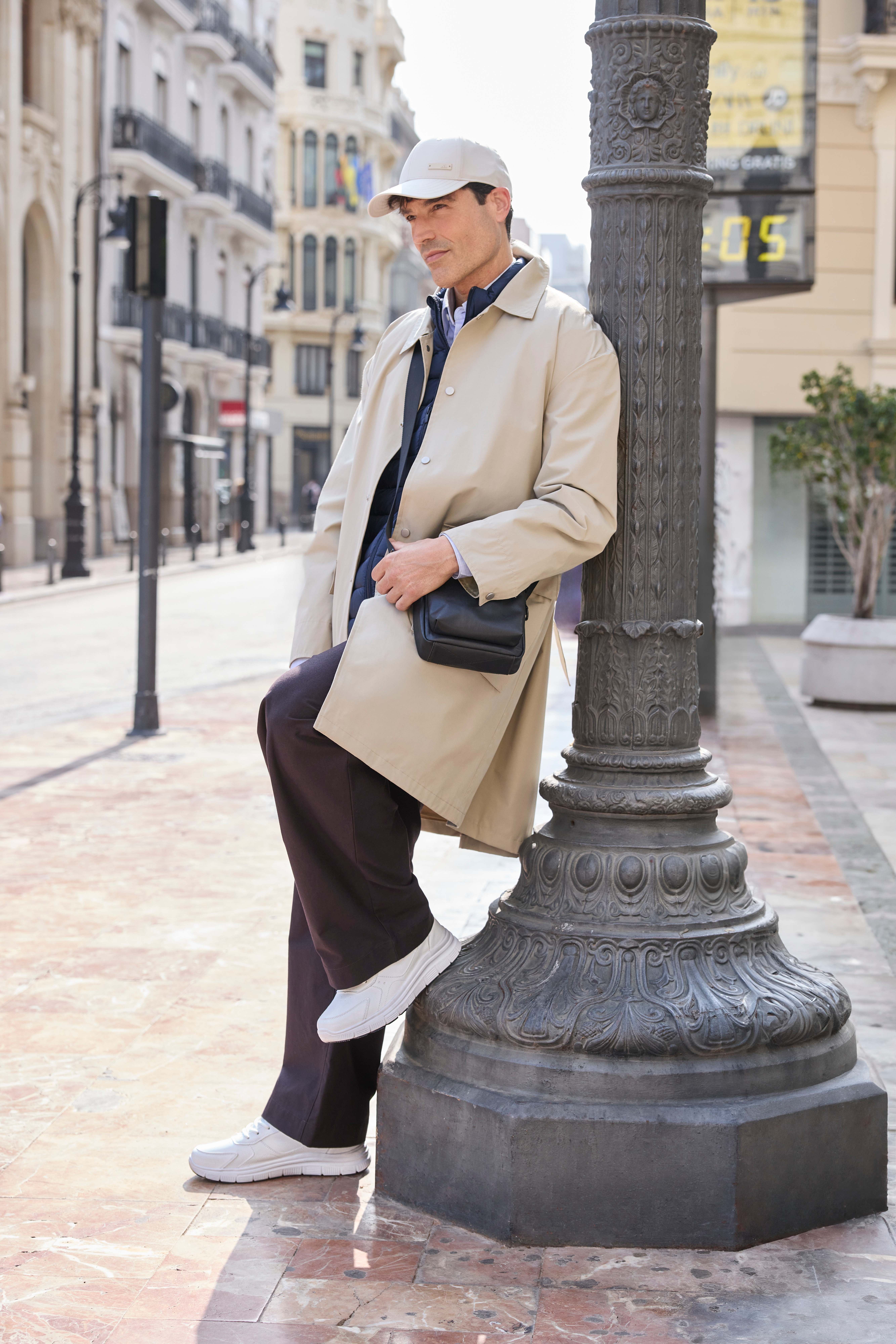 Mann in Corporate Core-Style, trägt beigen Trenchcoat mit dunkler Hose und weißen Sneakern, lehnt an Laterne