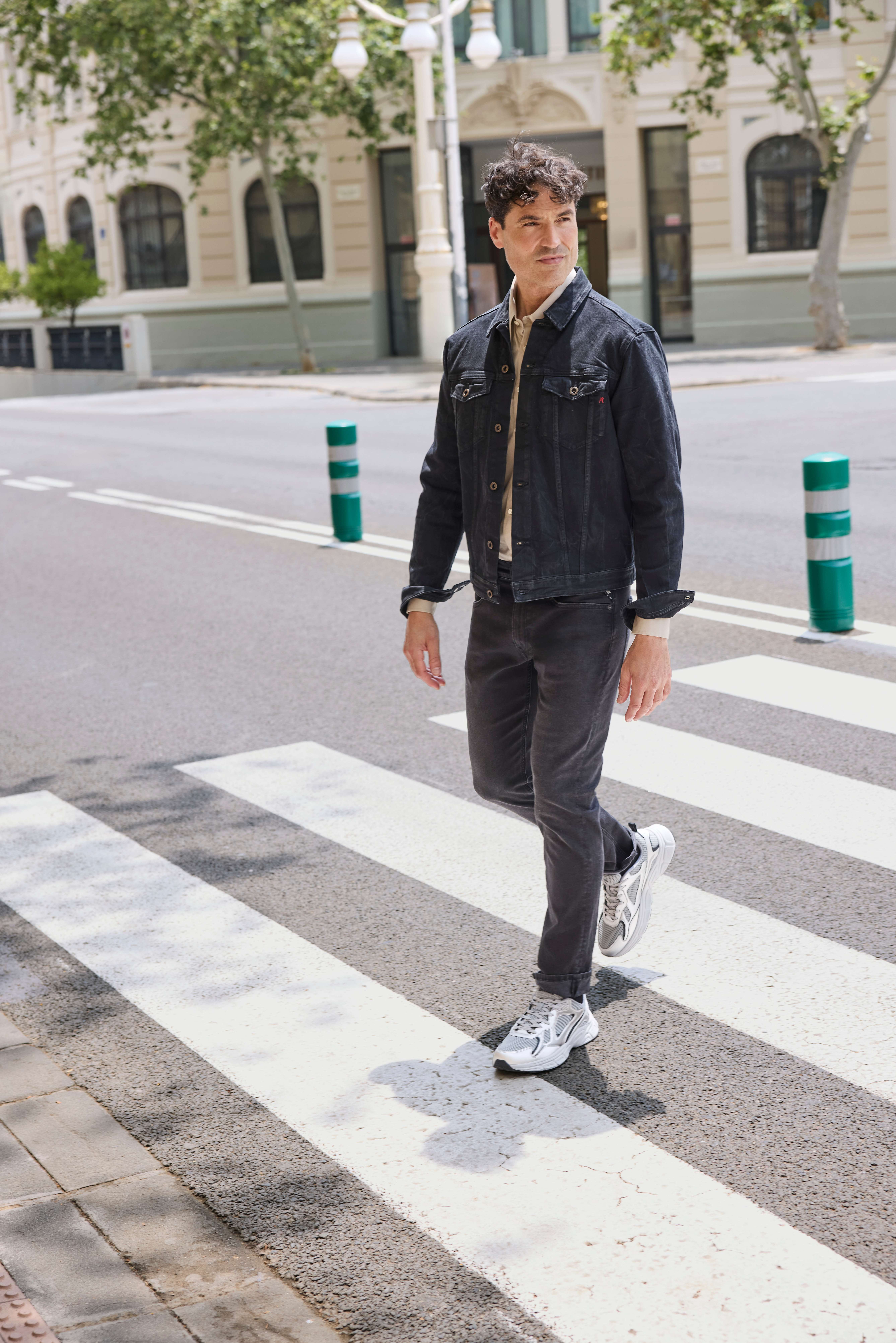 Ein Mann läuft auf einem Zebrastreifen und trägt eine schwarze Jeansjacke, ein beiges Hemd, eine schwarze Jeans und graue Sneakers