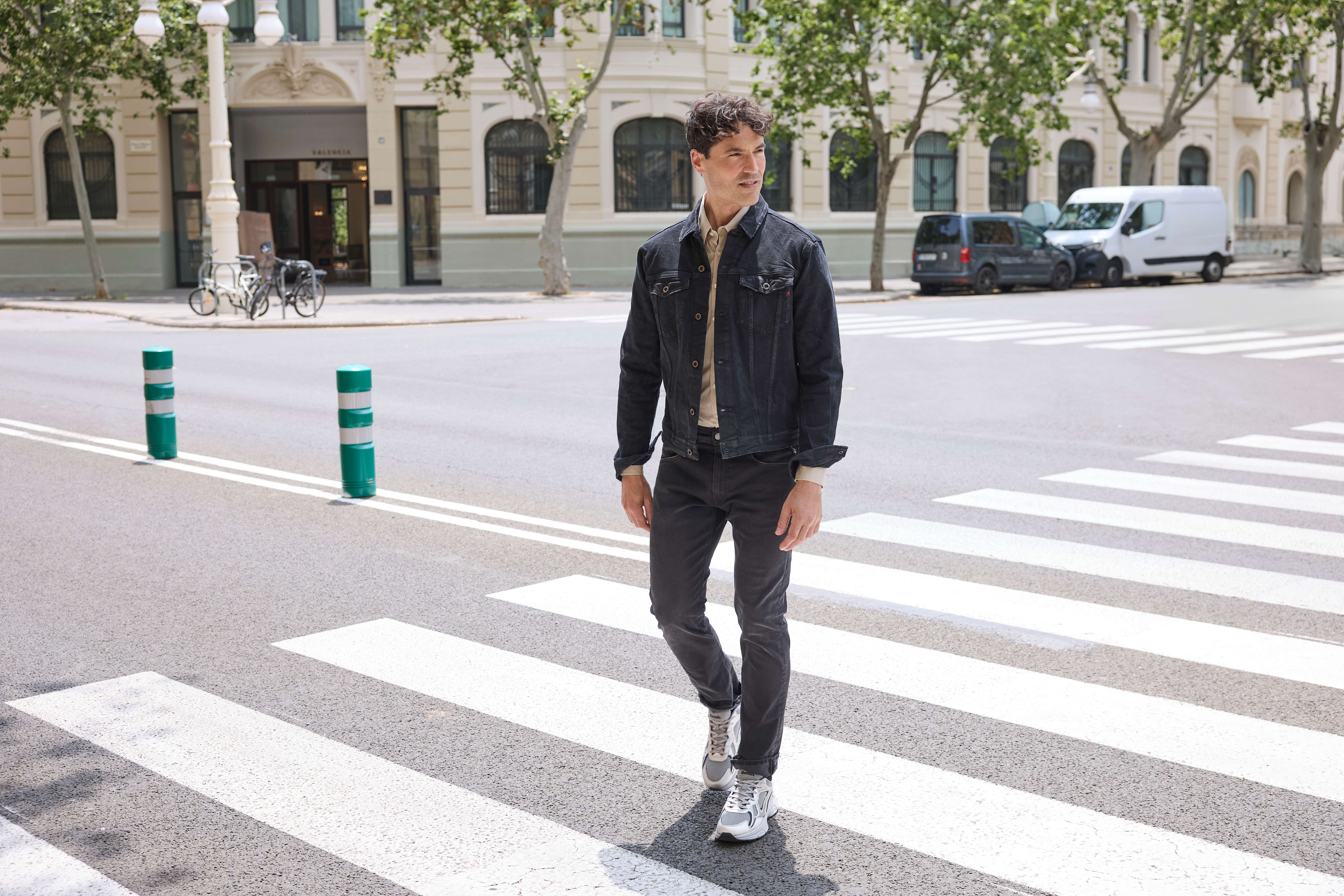 Ein Mann läuft auf einem Zebrastreifen und trägt eine schwarze Jeansjacke, ein beiges Hemd, eine schwarze Jeans und graue Sneakers