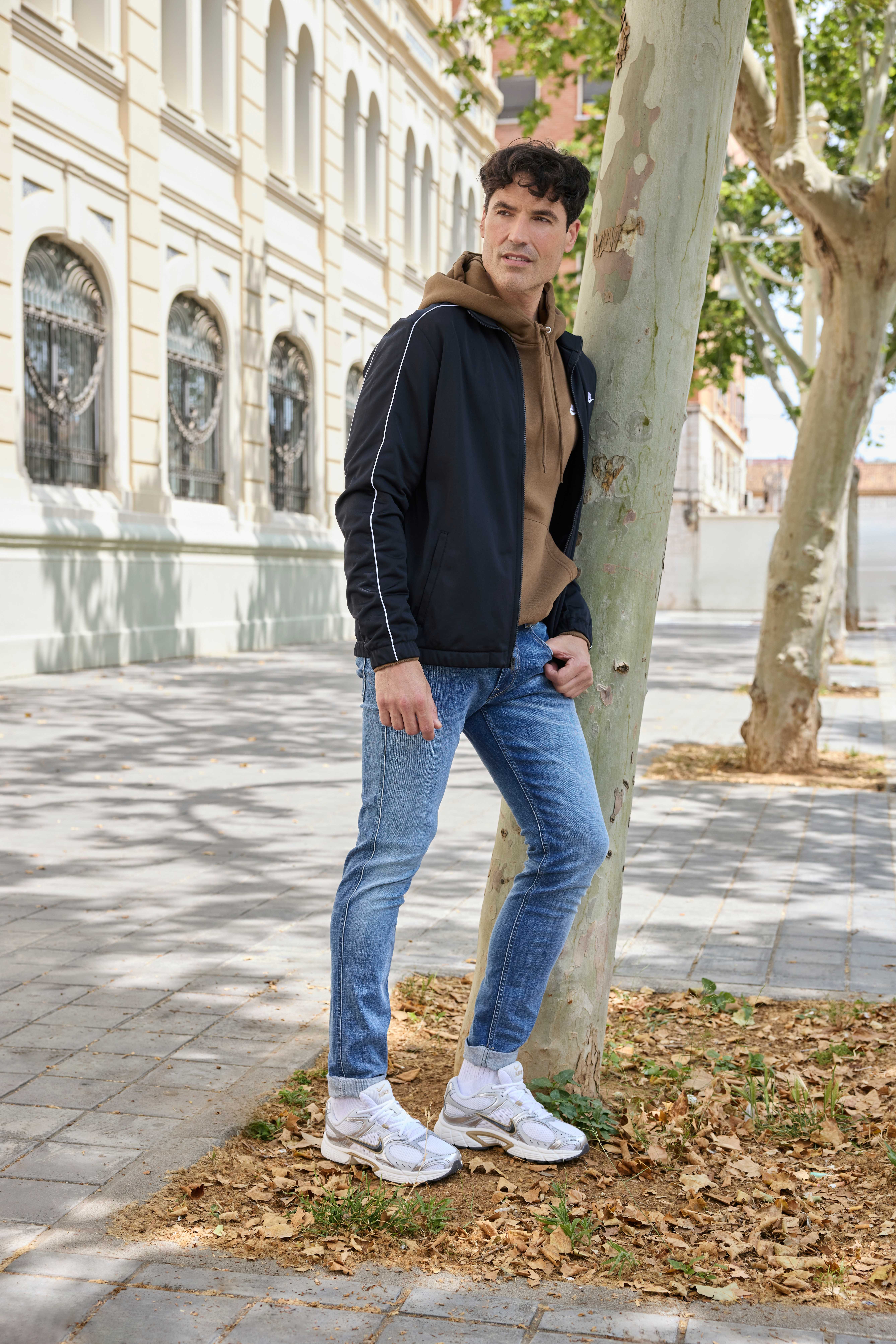 Mann mit schwarzem Track-Jacket und braunem Hoodie, lehnt an Baum