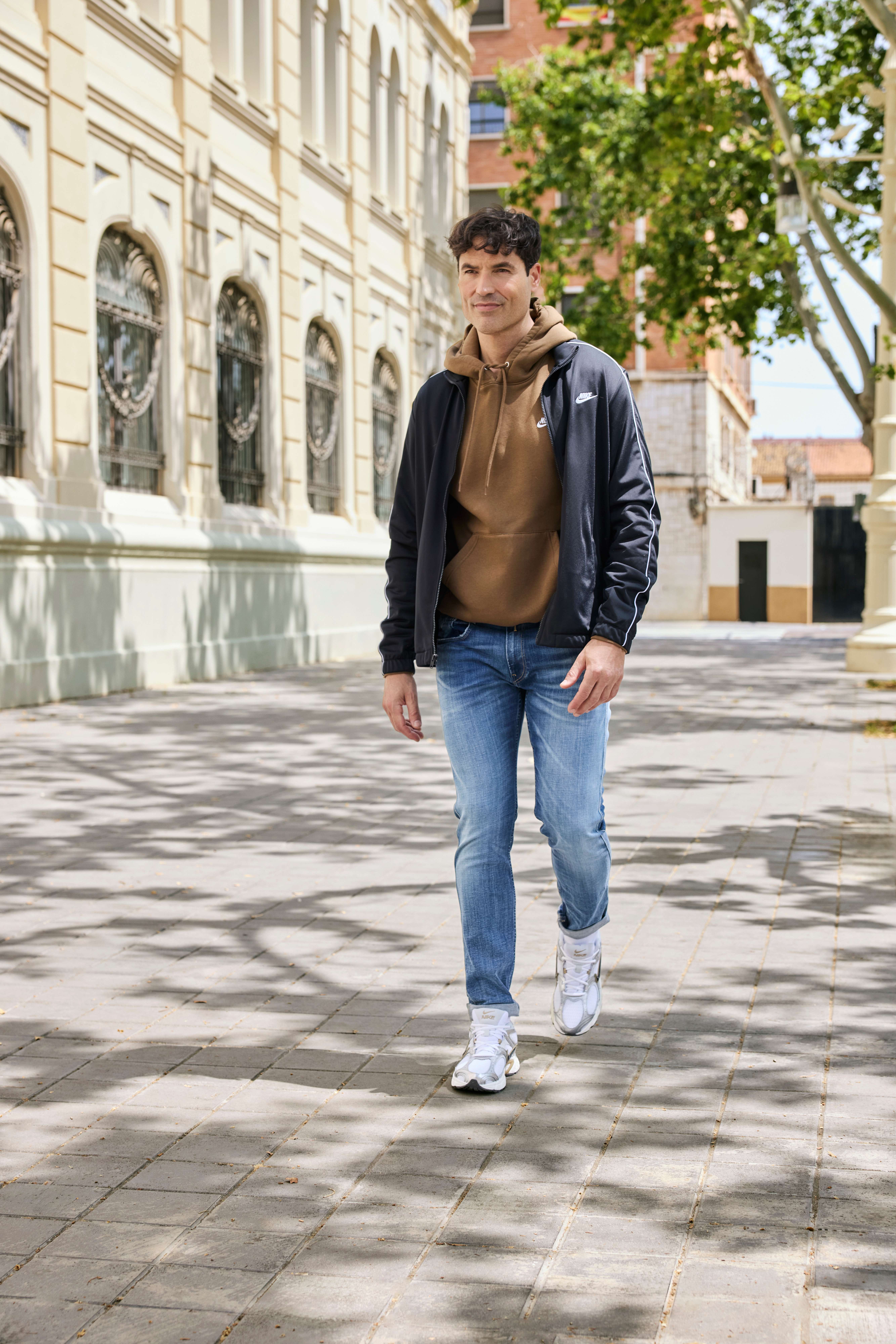 Ein Mann trägt eine schwarze Sportjacke, einen braunen Hoodie, eine blaue Jeans und weiße Sneakers