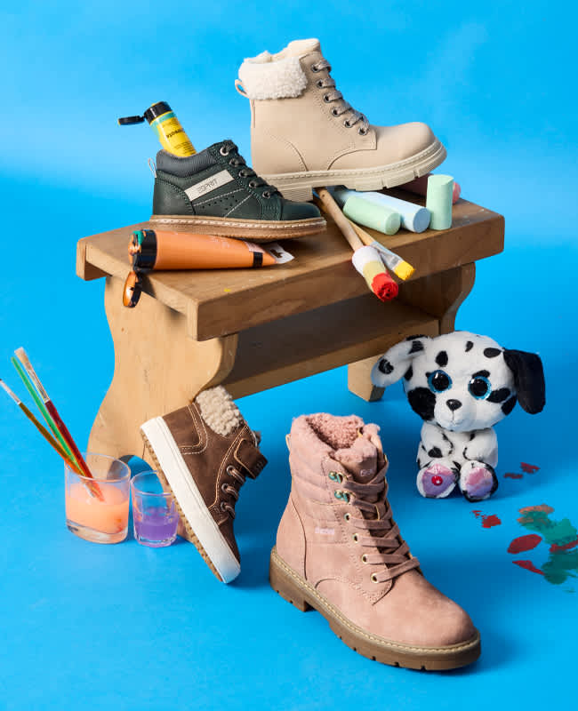Bunte Kinder-Schnürboots und Lauflerner auf und neben einer kleinen Holzbank, umgeben von Malutensilien, vor blauem Hintergrund.