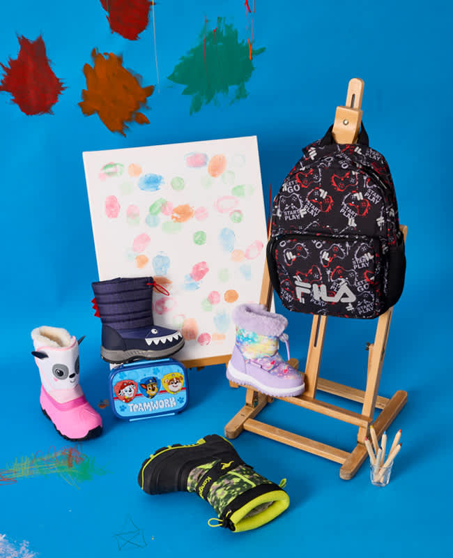 Bunte Kinder-Winterstiefel und ein FILA-Rucksack vor einer Staffelei, vor blauen Hintergrund.