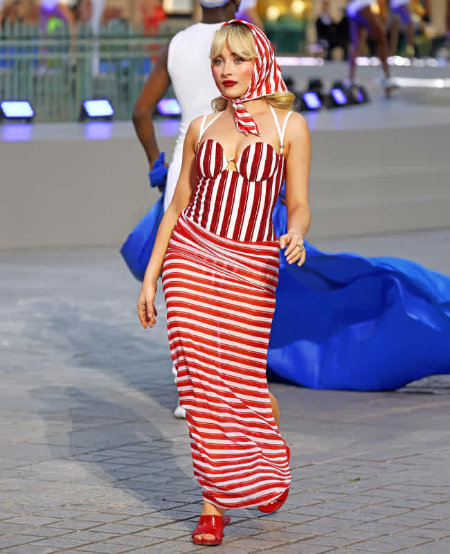 Schauspielerin Sabrina Carpenter im 60er-Jahre-Gesamt-Look, mit rot-weiß gestreiftem Korsett-Kleid samt passendem Kopftuch. Dazu trägt sie rote Sandalen.