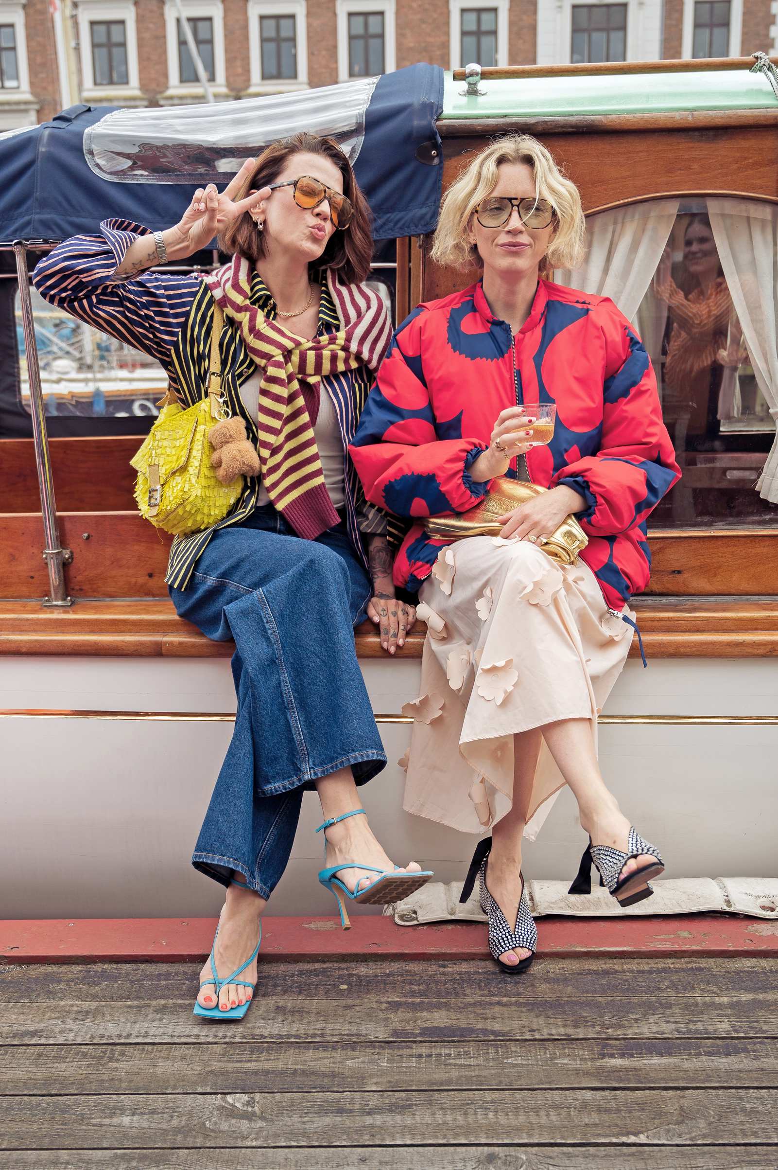 Zwei Damen tragen bunte Outfits für intensives Colour-Blocking, nebeneinander sitzend auf der Holzreling eines Boots