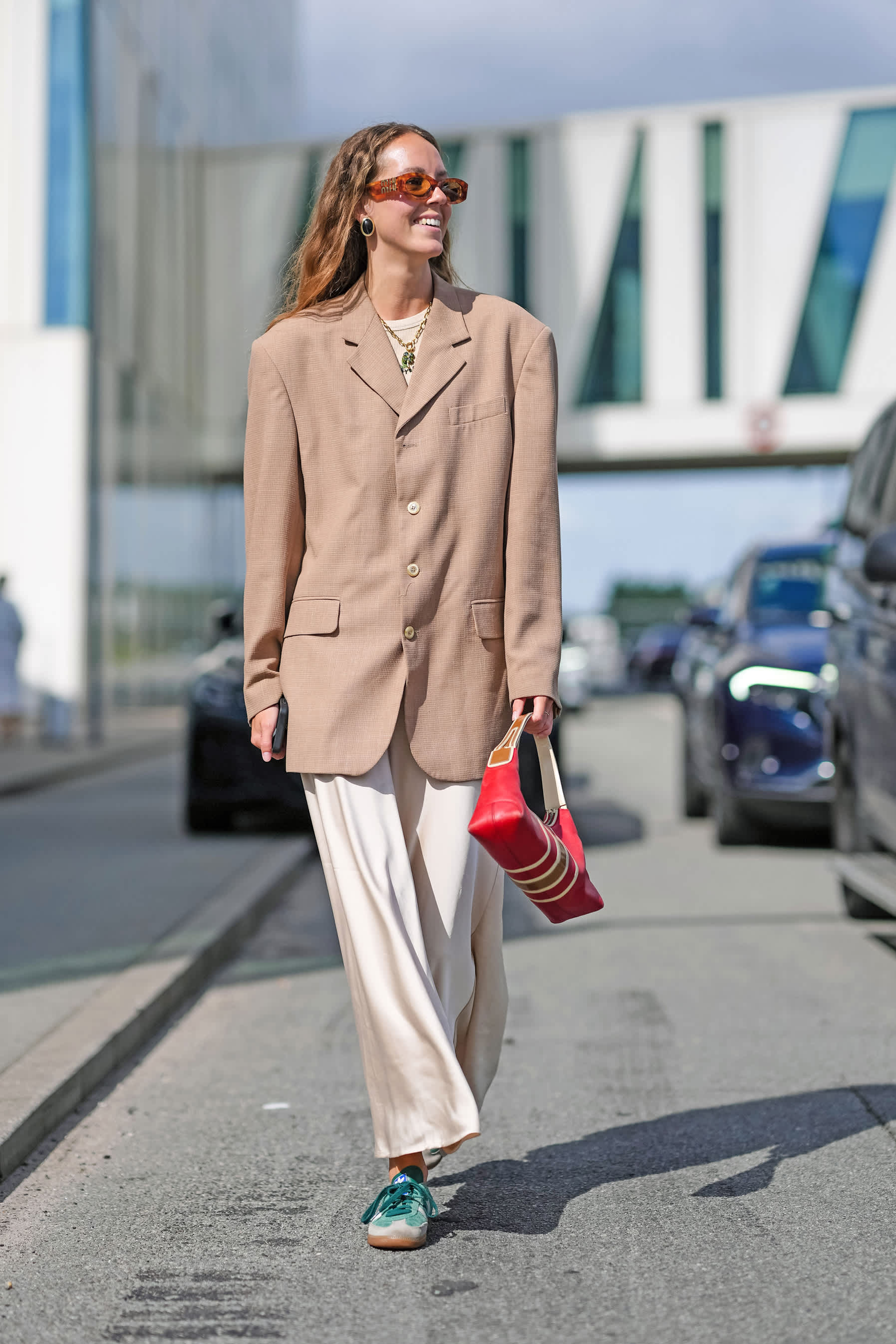 Frau in beigefarbenem Blazer, weißer Hose und grünen Sneakern, mit roter Handtasche in Hand, spazierend am Straßenrand