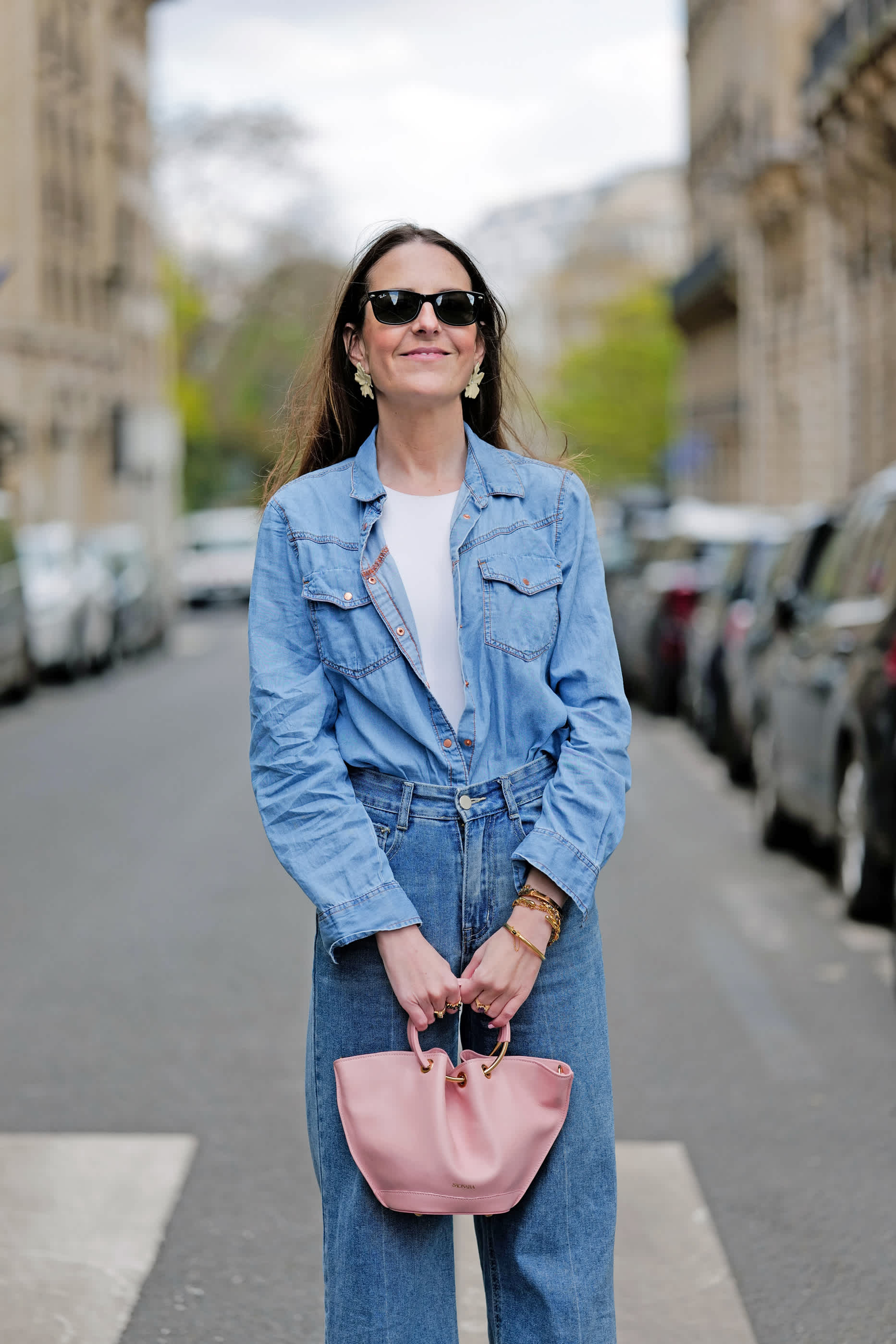 Frau trägt Jeanshemd und Jeanshose für einen Denim on Denim-Look und hält eine rosa Handtasche mit beiden Händen. Sie blickt selbstbewusst, Autos im Hintergrund