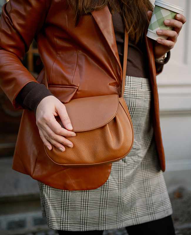 Nahaufnahme einer Person mit braunem Lederblazer und kariertem Rock, Hand liegt an brauner Handtasche