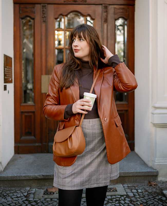 Frau in Lederblazer mit kariertem Rock, Tasche und Kaffeebecher vor einer Holztür, Outfit im Dark Academia Style