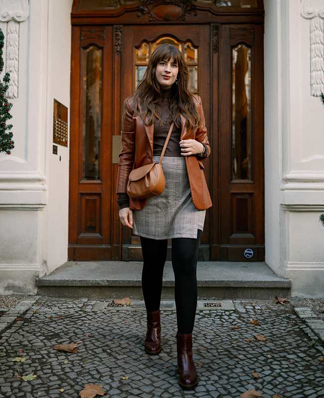 Frau in einem eleganten Herbst-Outfit mit Lederblazer, kariertem Rock und Stiefeletten, stehend vor einer Holztür.