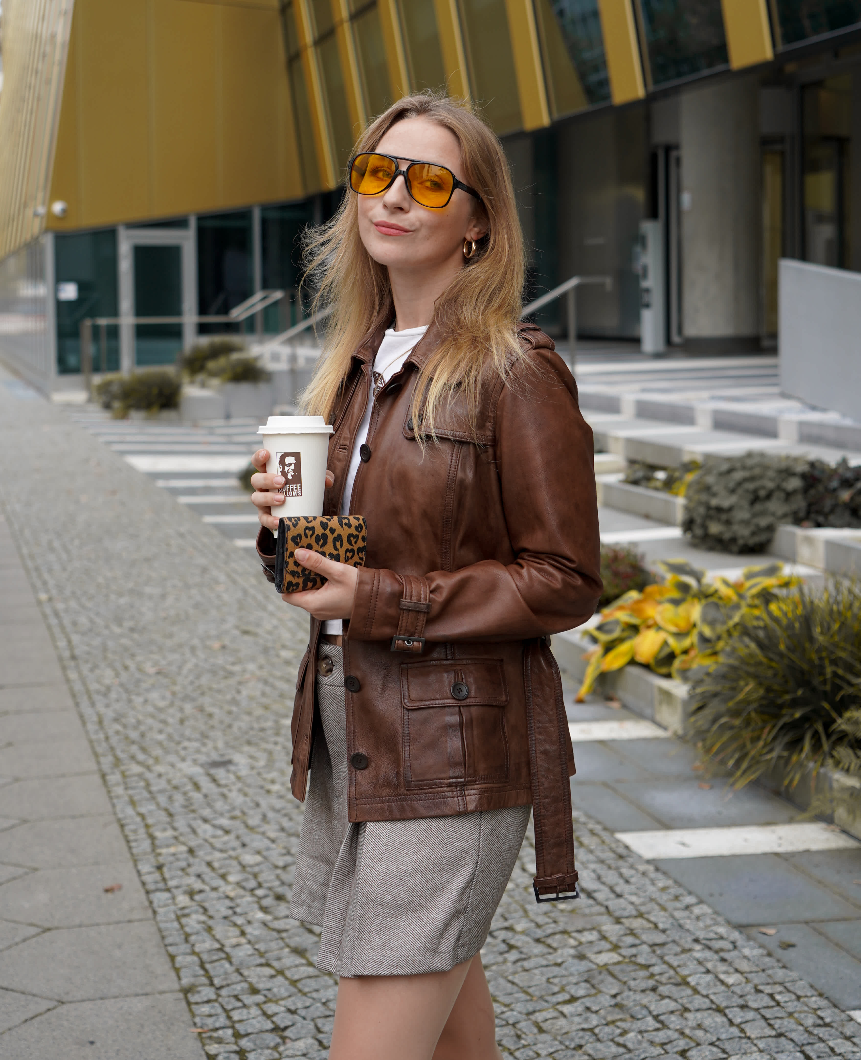 Frau mit brauner Lederjacke, Sonnenbrille und Coffee-to-go-Becher, lässig stehend vor urbanem Hintergrund