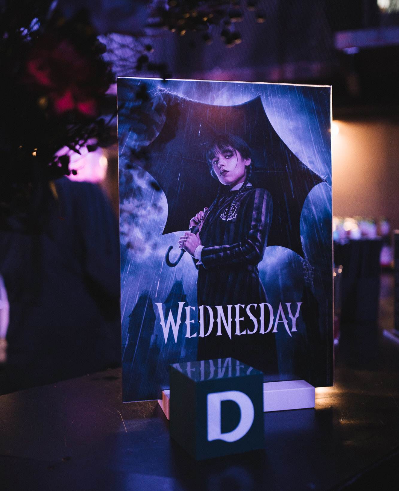 Promotional-Poster für die Serie "Wednesday" mit Abbildung der Protagonistin Wednesday Addams, davor steht ein kleiner grüner Würfel mit Deichmann-Logo