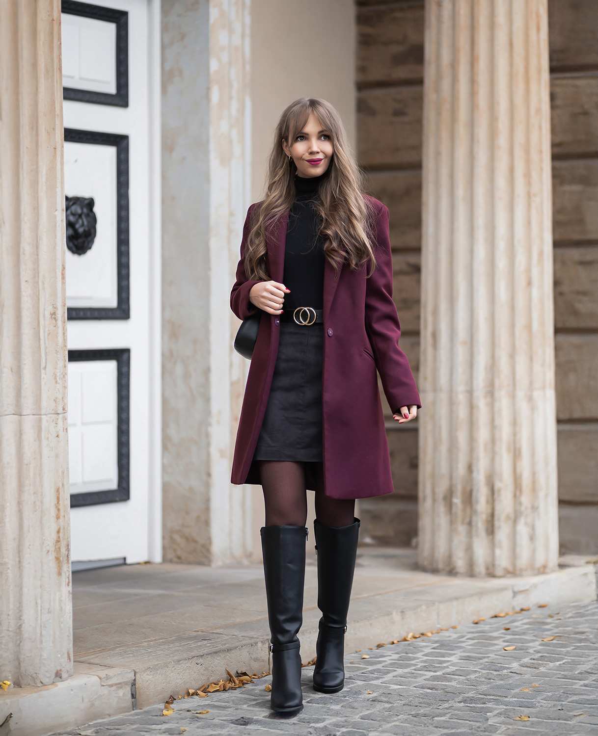Frau mit Outfit-Kombination In Burgunder und Schwarz, trägt roten Mantel, schwarzen Pullover, Rock und Stiefel zusammen mit schwarzer Tasche