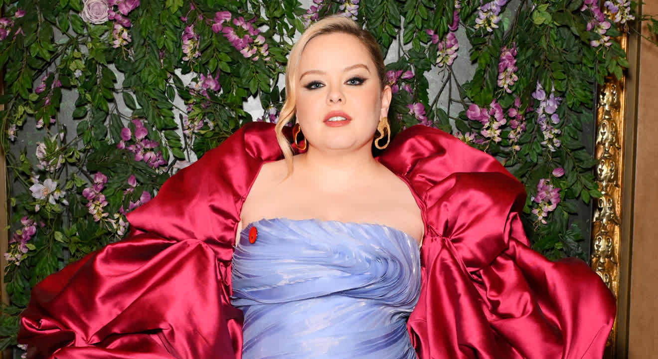 Schauspielerin Nicola Coughlan steht in lavendelblauem Kleid mit rotem Umhang vor einer Blumenwand. 