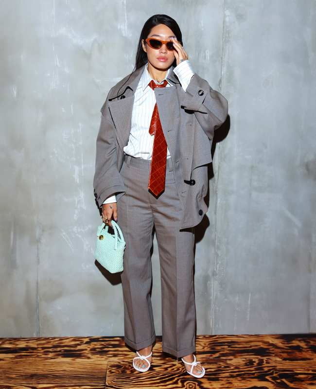 Designerin Peggy Gou in übergroßem grauen Anzug mit gestreiftem Hemd als Ton-in-Ton-Look, roter Krawatte und einer kleinen mintfarbenen Tasche, stehend vor einer Betonwand.