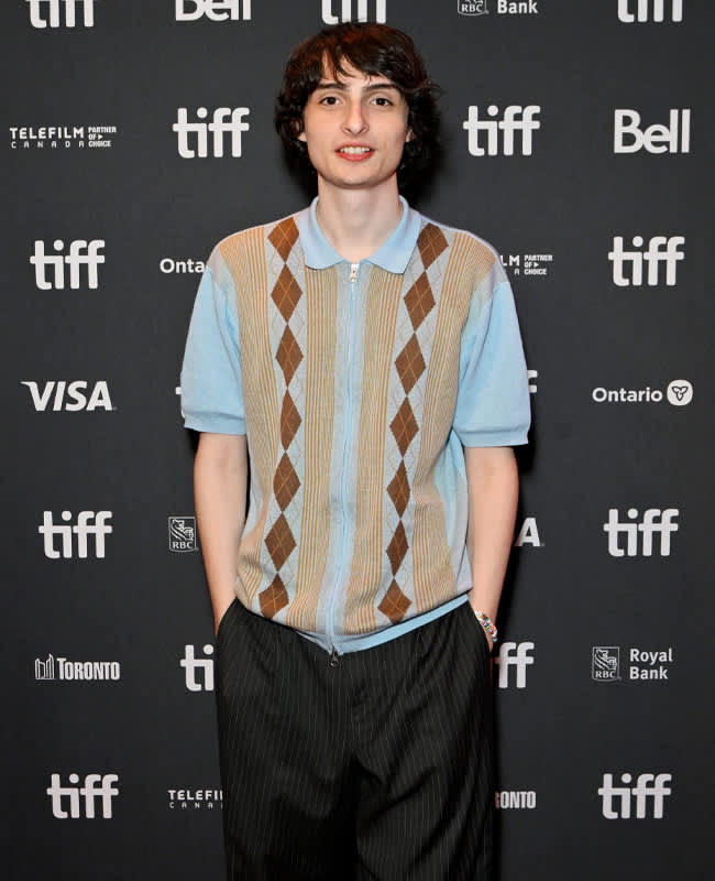 Schauspieler Finn Wolfhard in hellblauem Polohemd mit Rautenmuster und dunkler Stoffhose, stehend vor einer schwarzen Logo-Wand.