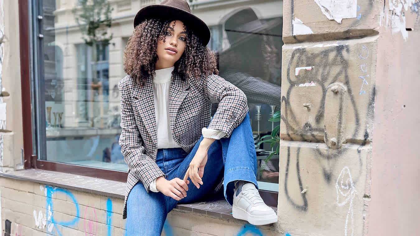 Frau mit Hut, kariertem Blazer mit Jeans und weißen Sneakern, sitzend auf einer Fensterbank.