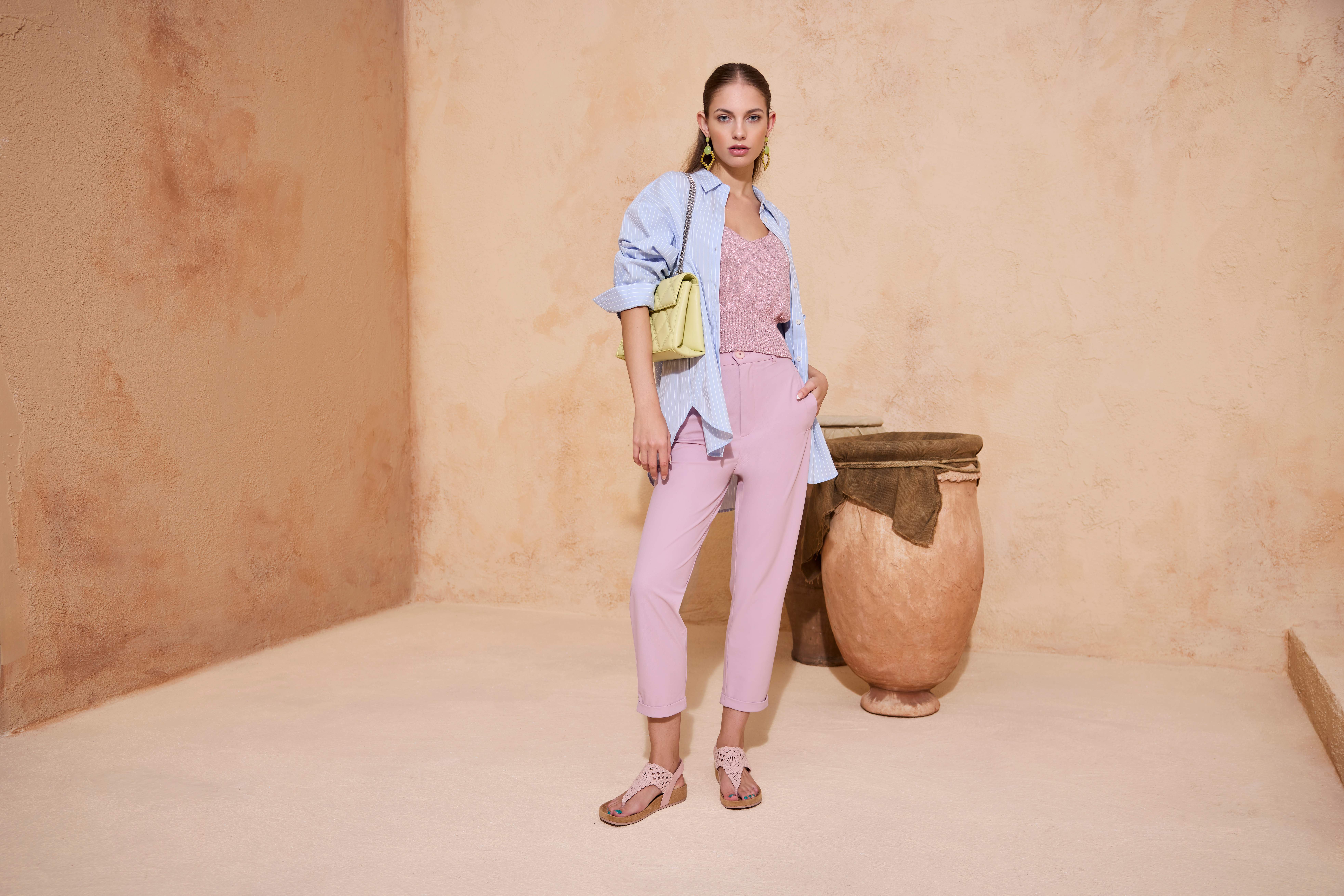Frau vor beigem Steinhintergrund, Rosa Hose, Rose Oberteil mit blauer Bluse, gelbe Handtasche