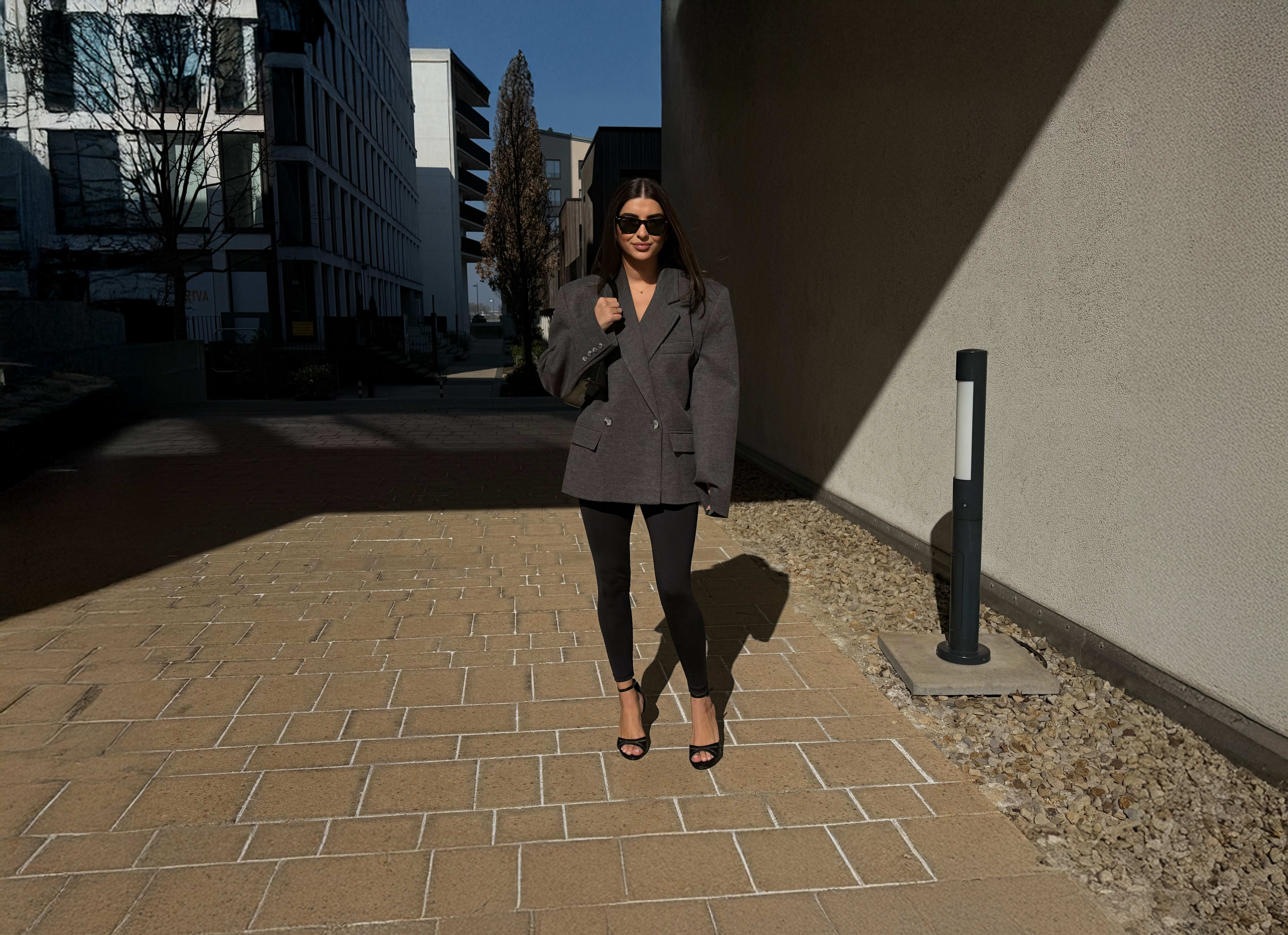 Frau mit schwarzer Leggins, Sandaletten und grauem Blazer mit passender Tasche zwischen zwei Häusern