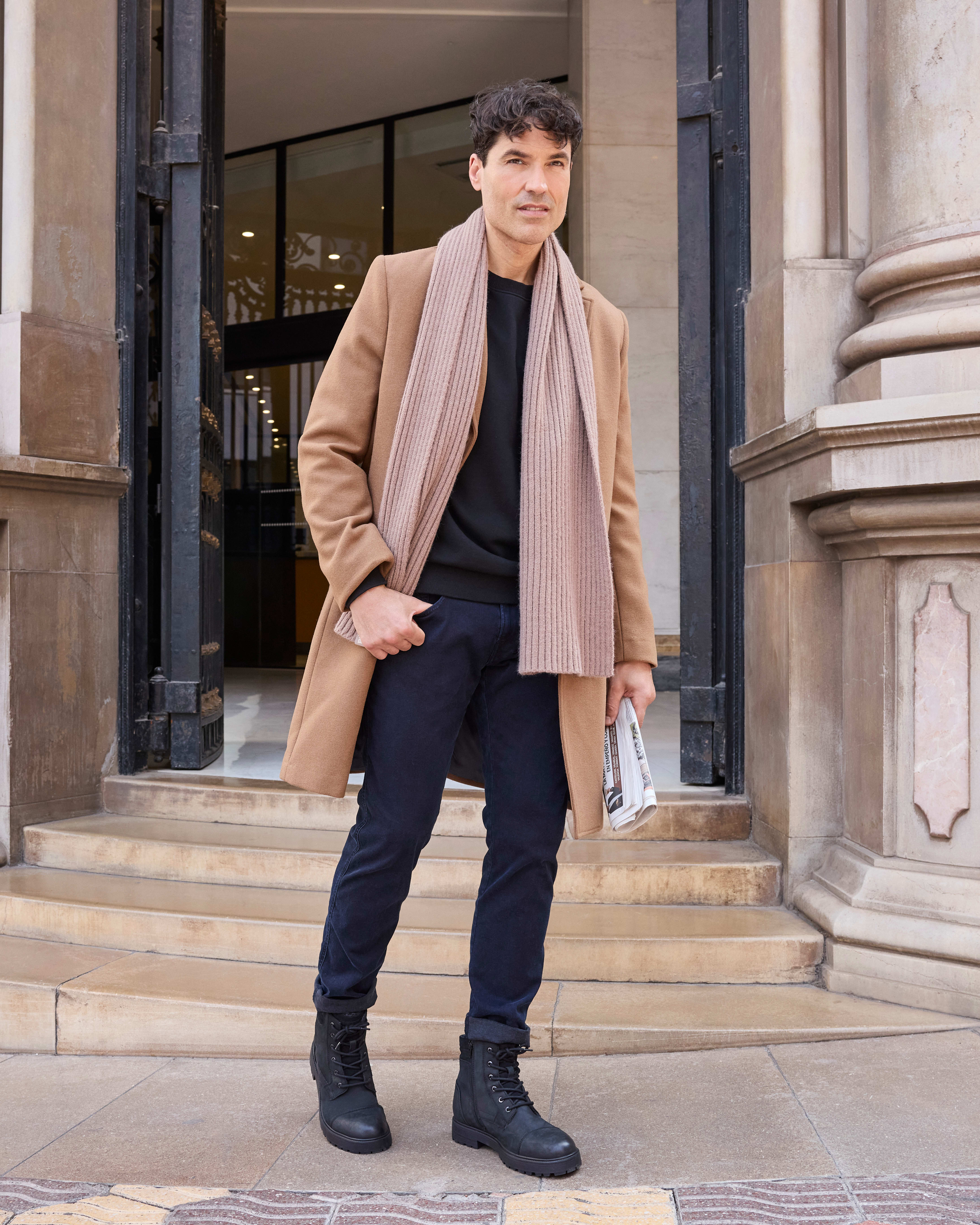 Ein Mann trägt einen beigen Mantel, einen Schal, ein schwarzes Shirt und eine schwarze Jeans