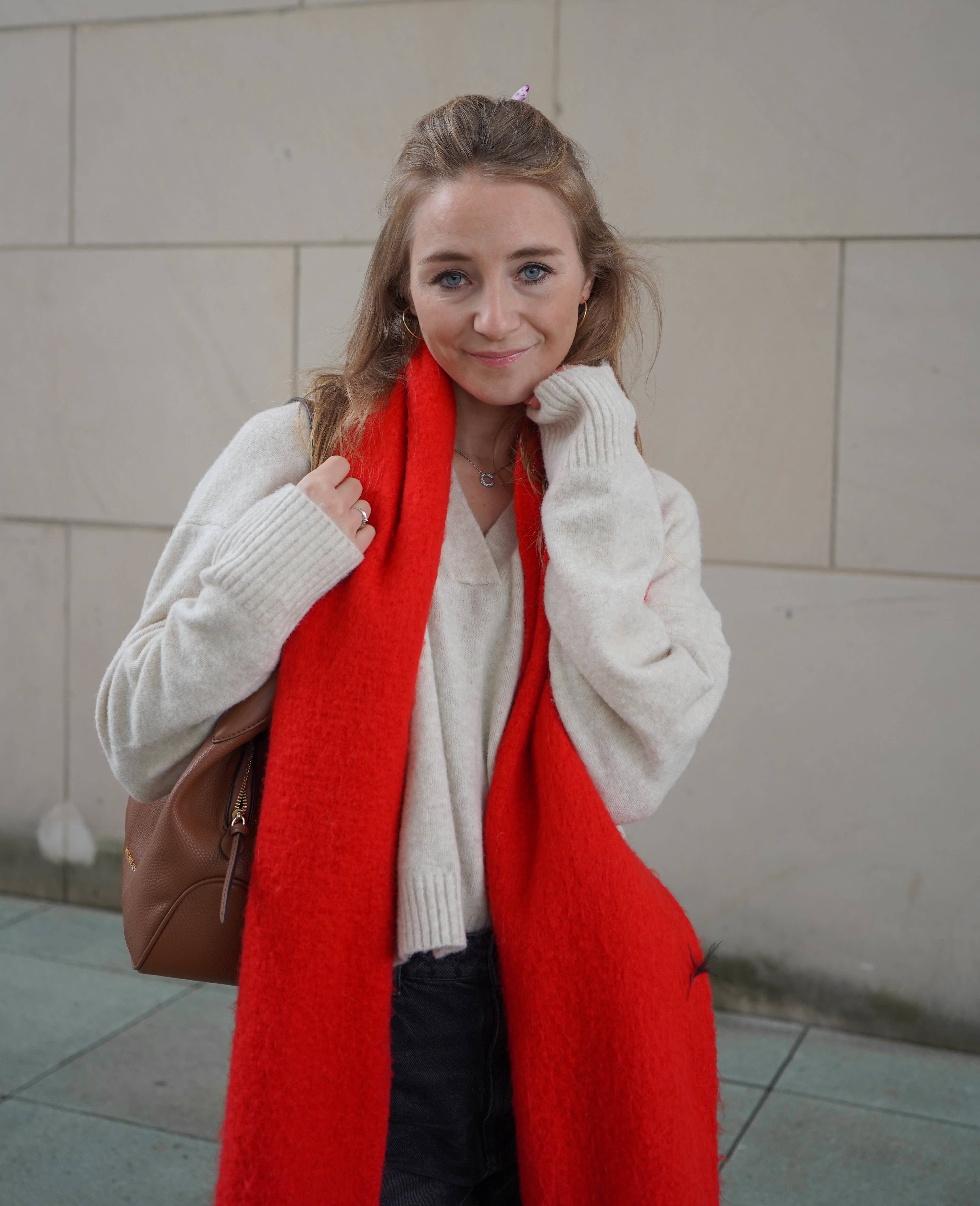 Frau in Cozy-Outfit mit rotem Schal, weißem Pullover und brauner Tasche, Steinwand im Hintergrund