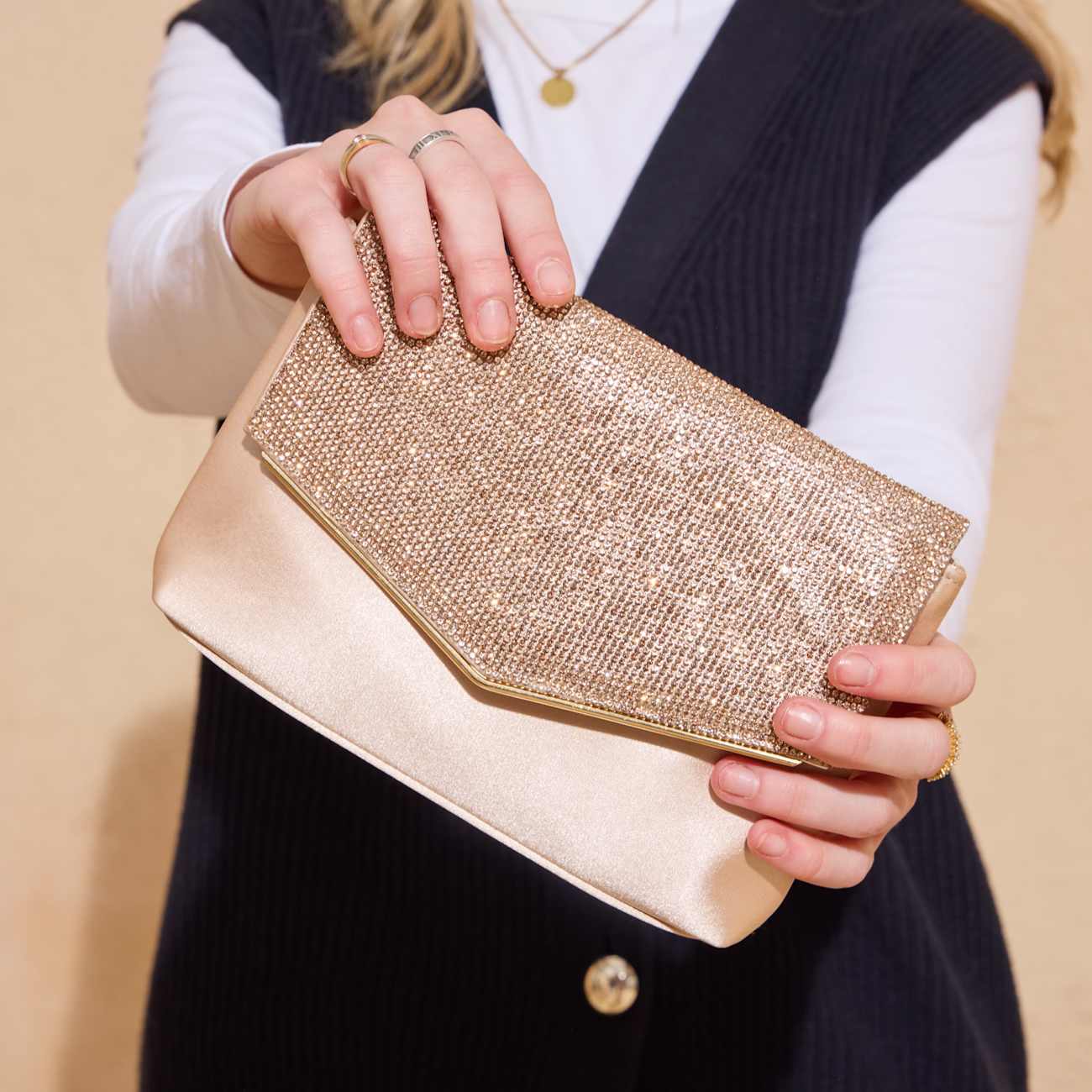 Person in schwarz-weißem Outfit hält mit beiden Händen eine schlichte, gold-schimmernde Glitzertasche 