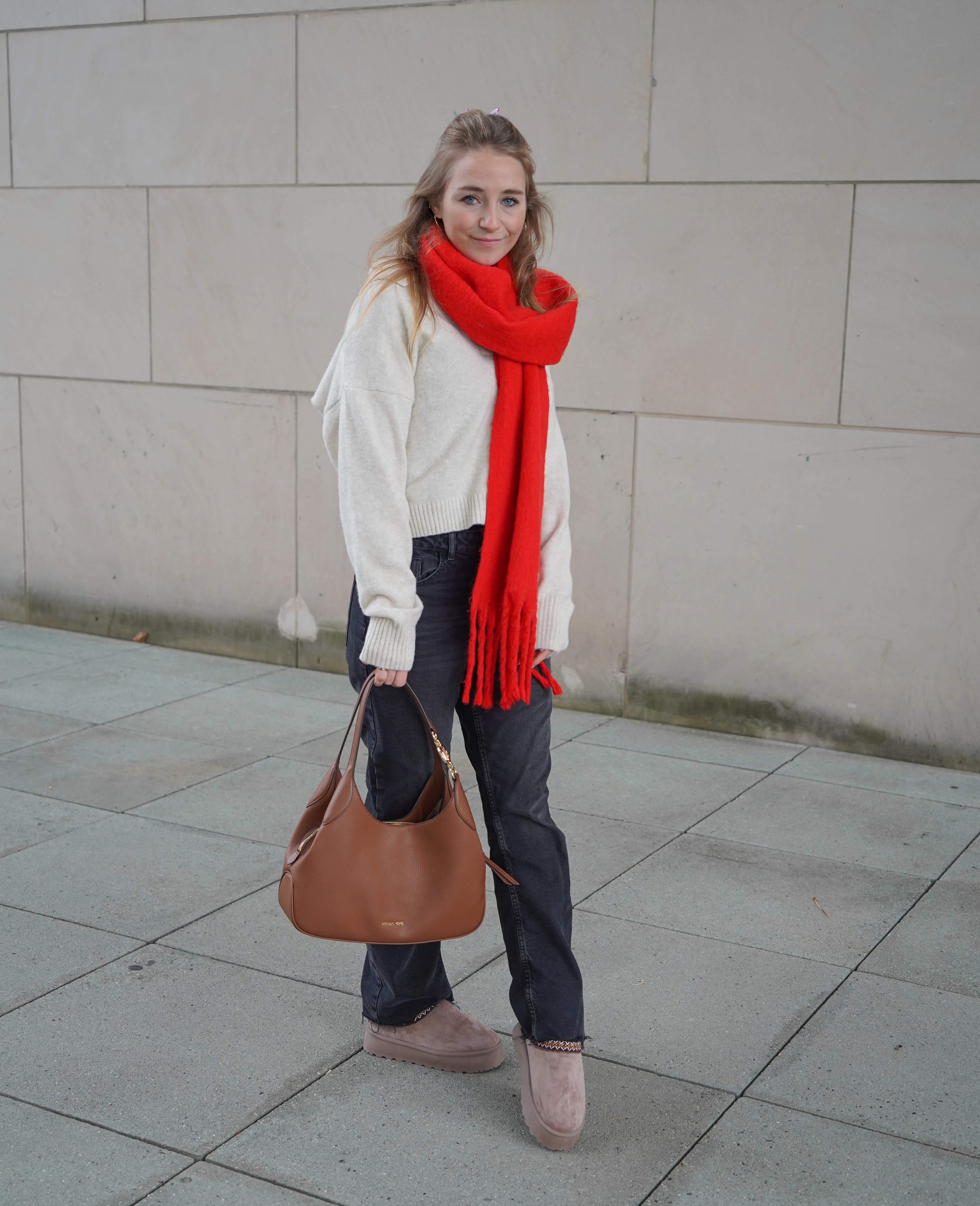 Frau trägt cozy Oversized-Pullover, kombiniert mit dickem, roten Schal, lässiger Jeans und beigen Pantoletten