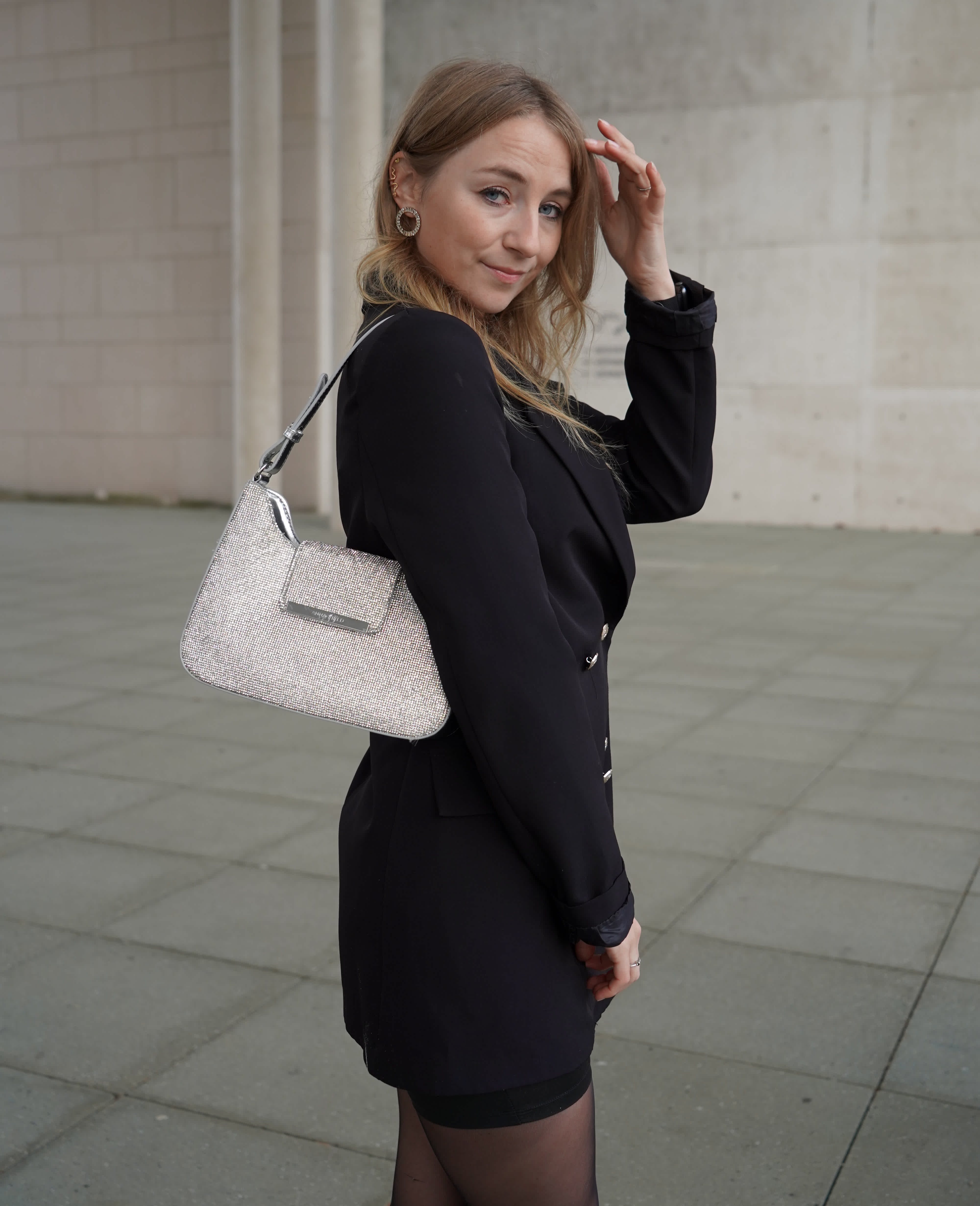 Frau trägt schwarzes Blazer-Kleid, kombiniert mit silberner Schultertasche und minimalistischen Accessoires, Blick seitlich zur Kamera