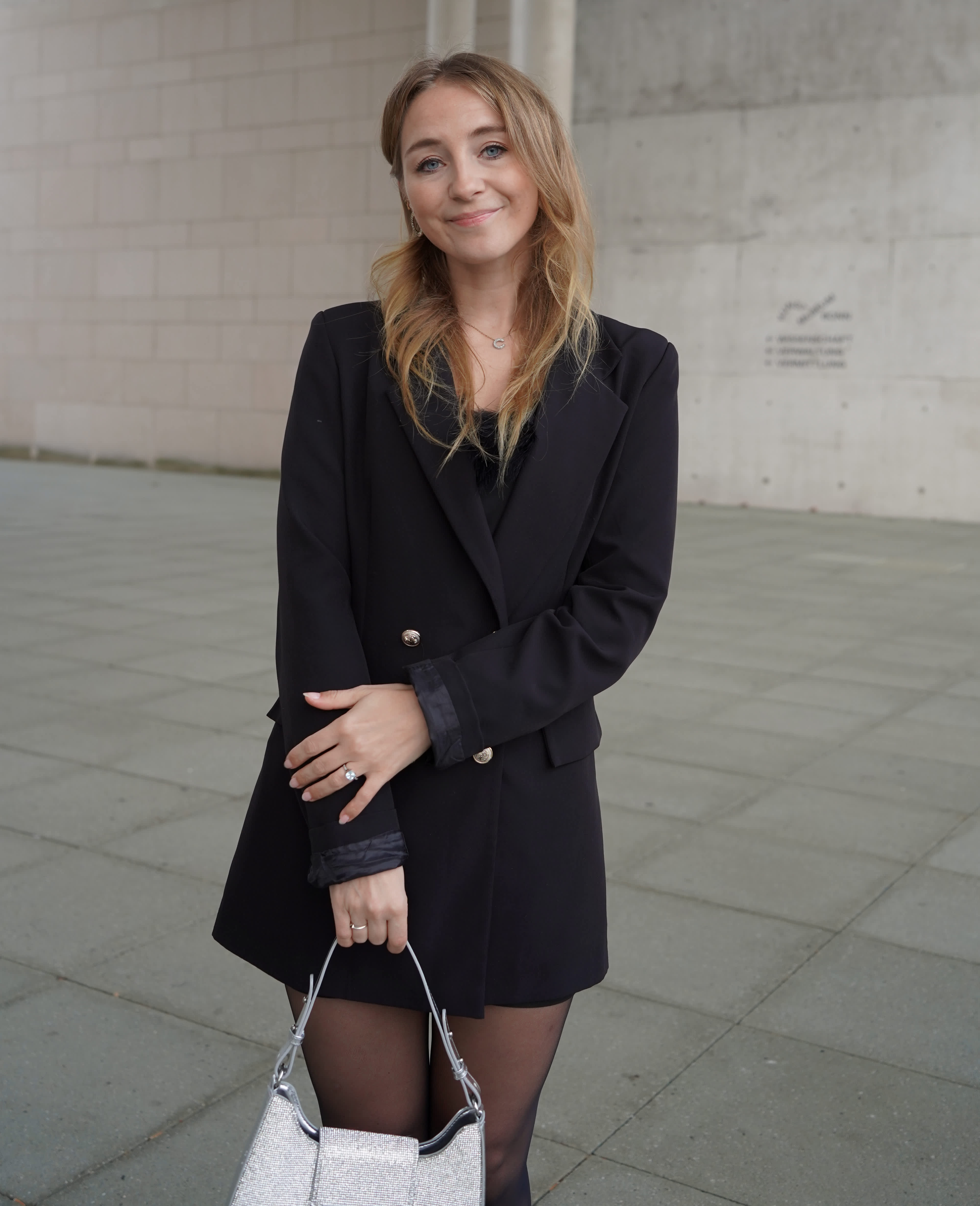 Frau in schwarzem Blazer-Dress, trägt silbern-glitzernde Mini-Handtasche, blickt lächelnd zur Kamera, vor urbaner Kulisse
