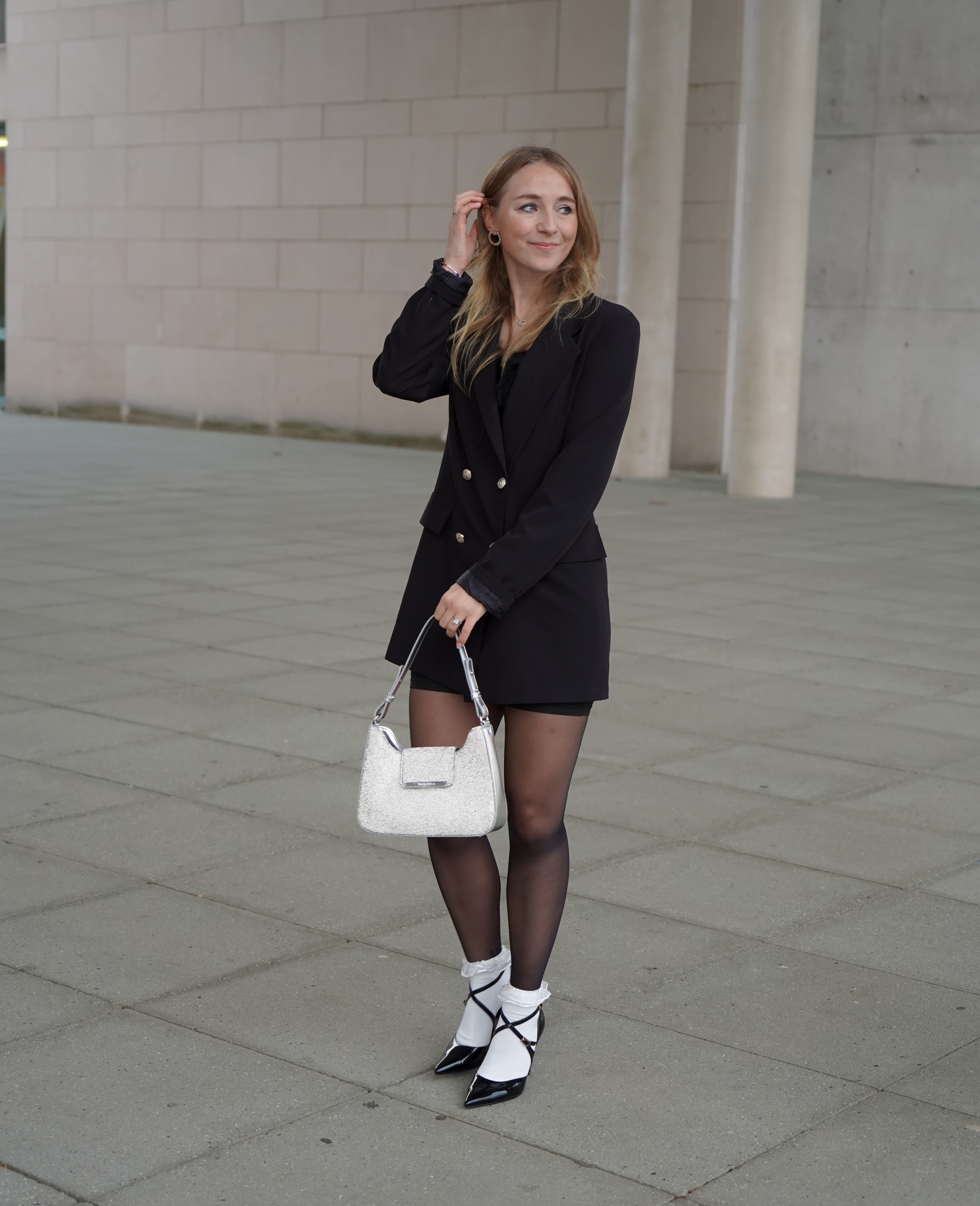 Frau in schwarzem Blazer-Mini-Dress mit schwarzen Lackpumps, trägt eine silbern-glitzernde Handtasche 