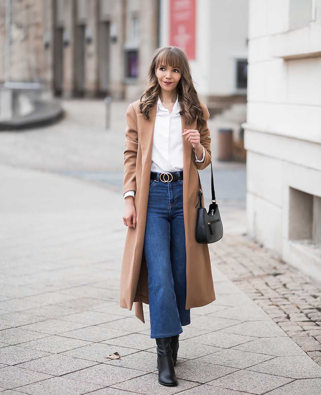 Frau trägt Camel Coat in Kombination mit blauer Jeanshose, weißer Bluse und schlichter Handtasche in Schwarz