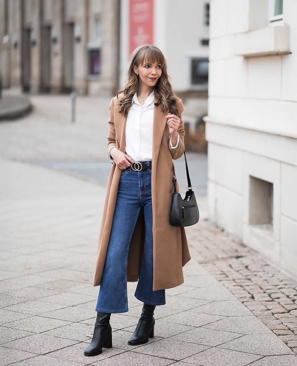 Frau in camel-farbenem Mantel, mit weißer Bluse und blauer High-Waist-Jeans mit weitem Bein, trägt Handtasche am Arm, vor urbaner Kulisse 