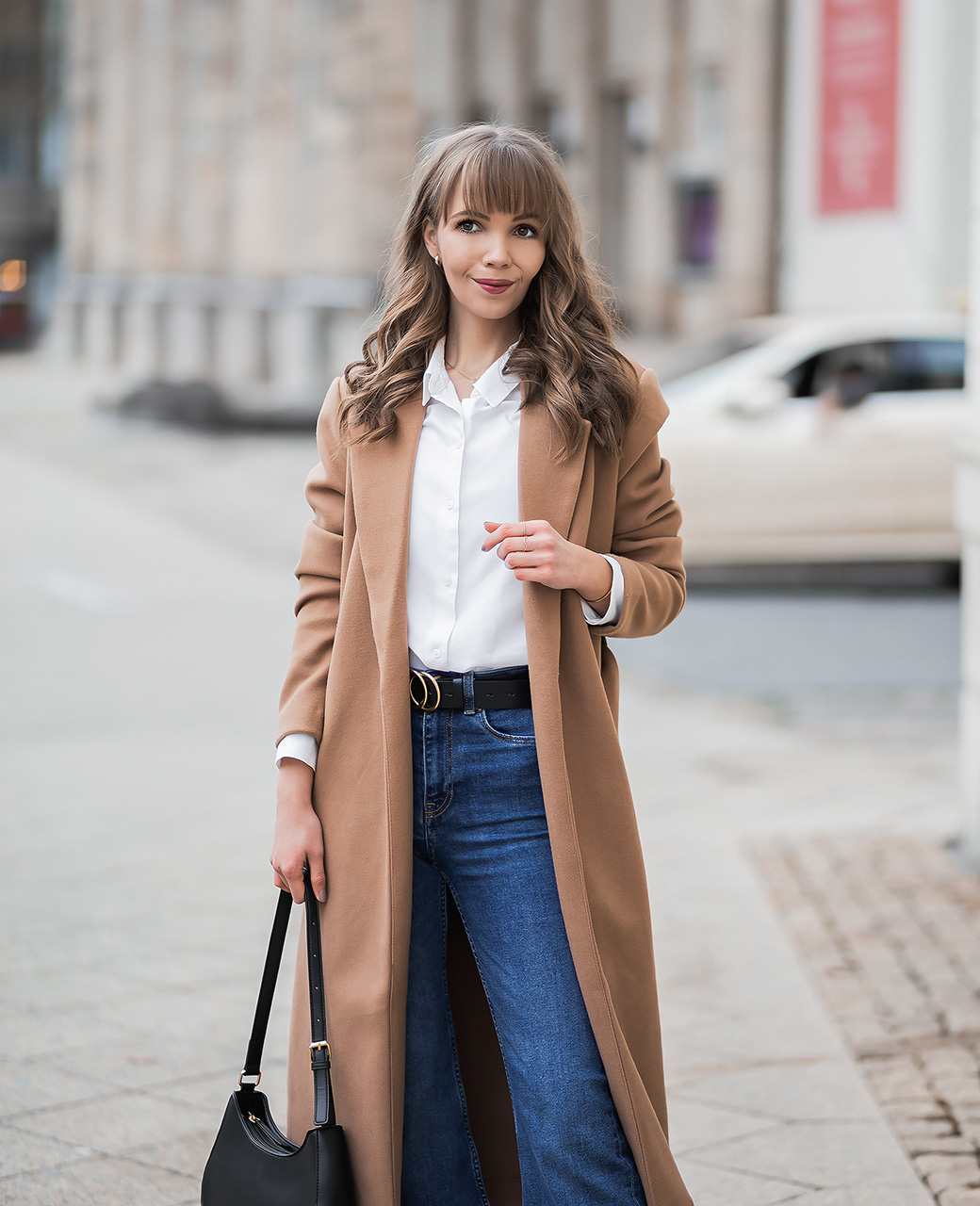 Frau in Camel Coat, kombiniert mit weißer Bluse und blauer High-Waist-Jeans, vor urbaner Kulisse