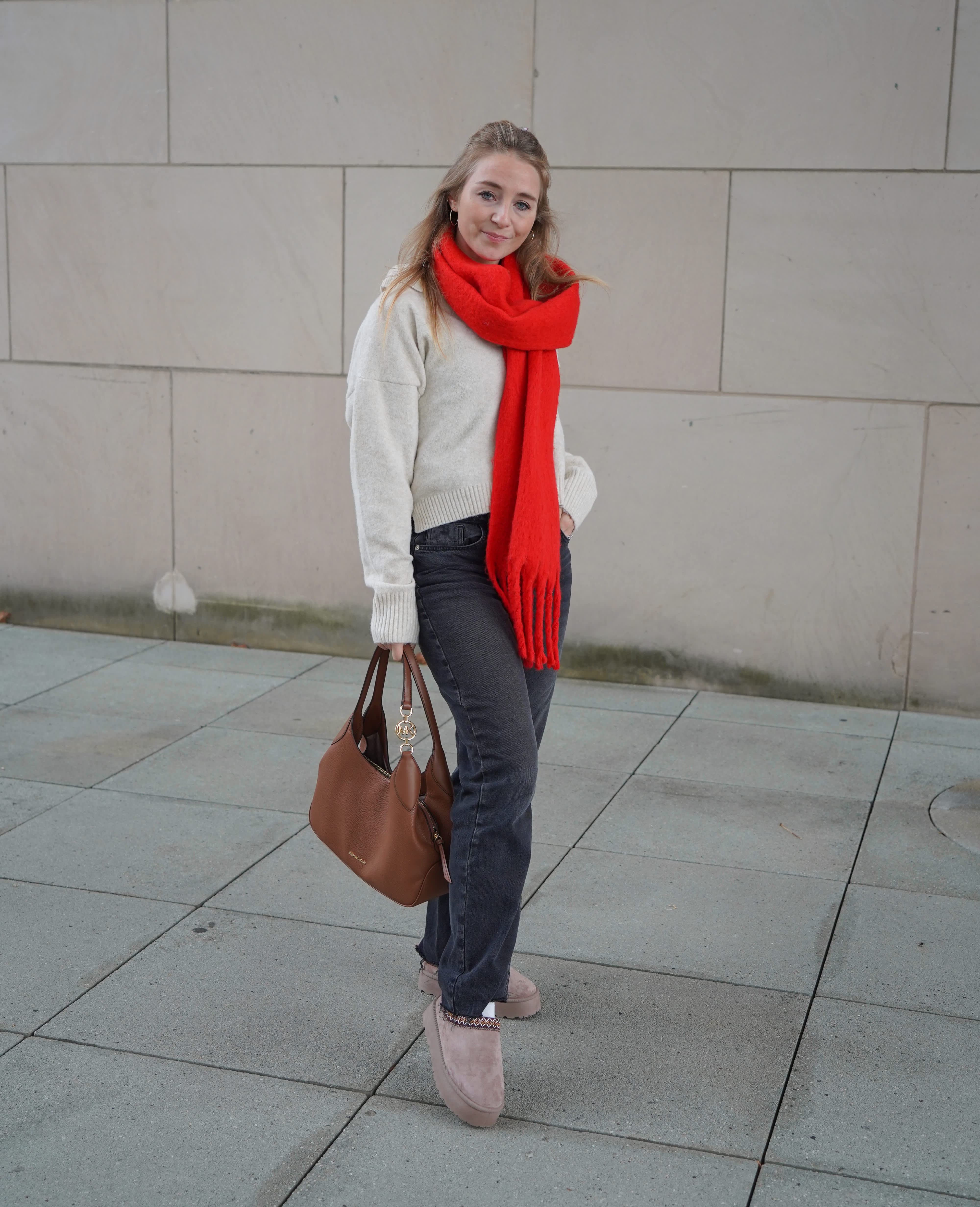 Frau in gemütlichem Oversized-Strickpullover, kombiniert mit Jeans, großem roten Schal und beigefarbenen Pantoletten