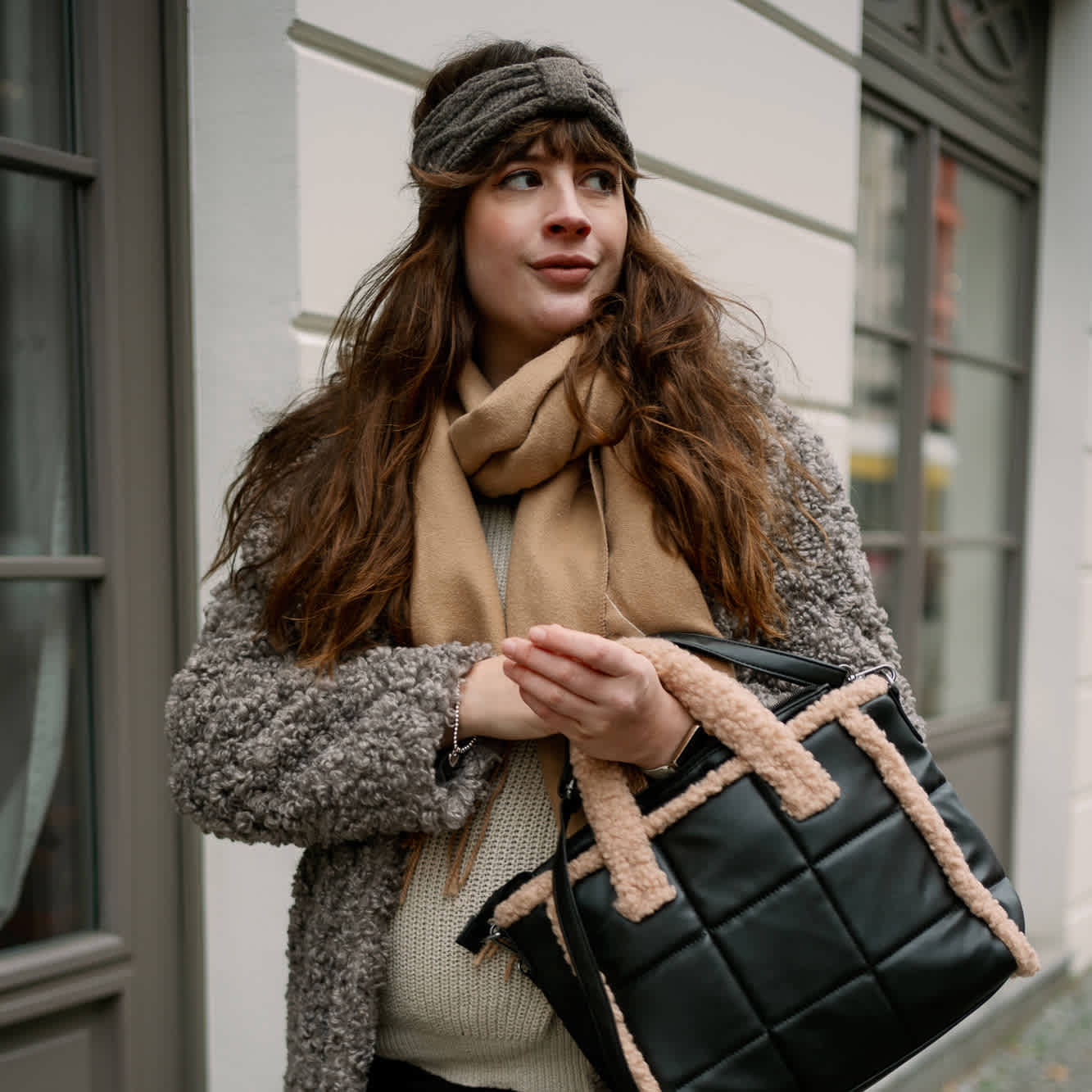 Frau in warmer Kleidung mit grauem Mantel und Schal in Beige, trägt eine Handtasche, vor urbaner Kulisse