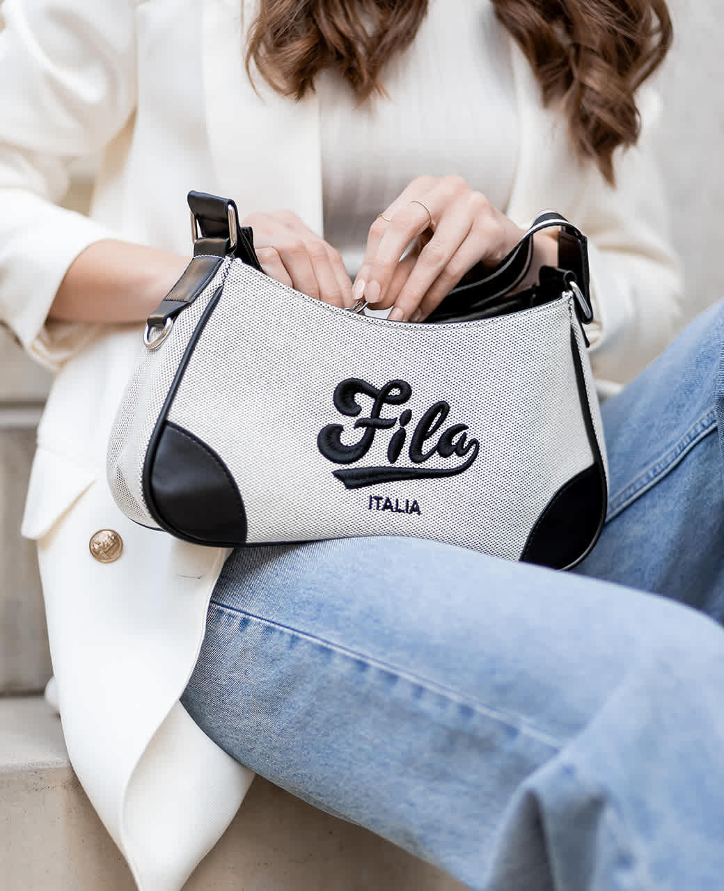 Frau mit weißem Oberteil trägt weiße Fila-Handtasche mit Schriftzug auf dem Schoß