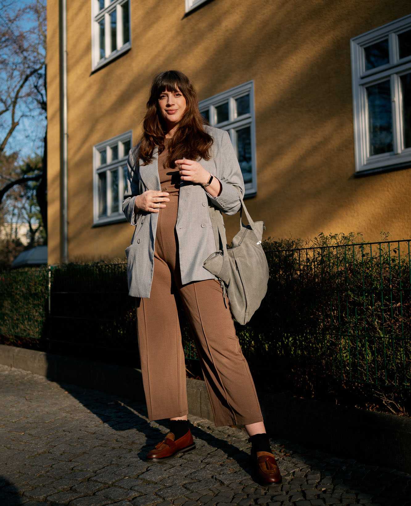 Frau mit braunen Jumpsuit und grauem Blazer vor gelber Häuserwand