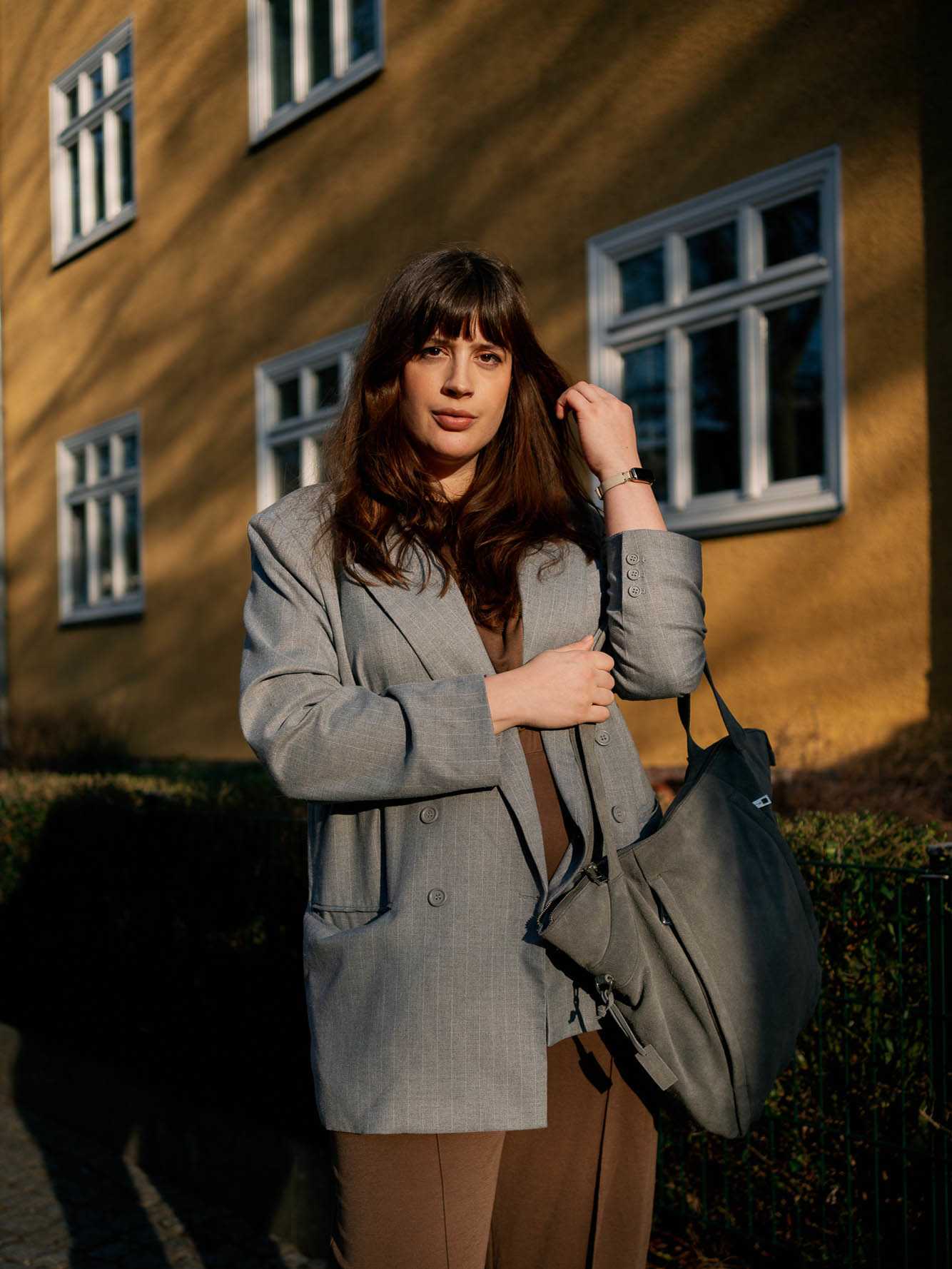 Frau mit braunen Jumpsuit und grauem Blazer vor gelber Häuserwand
