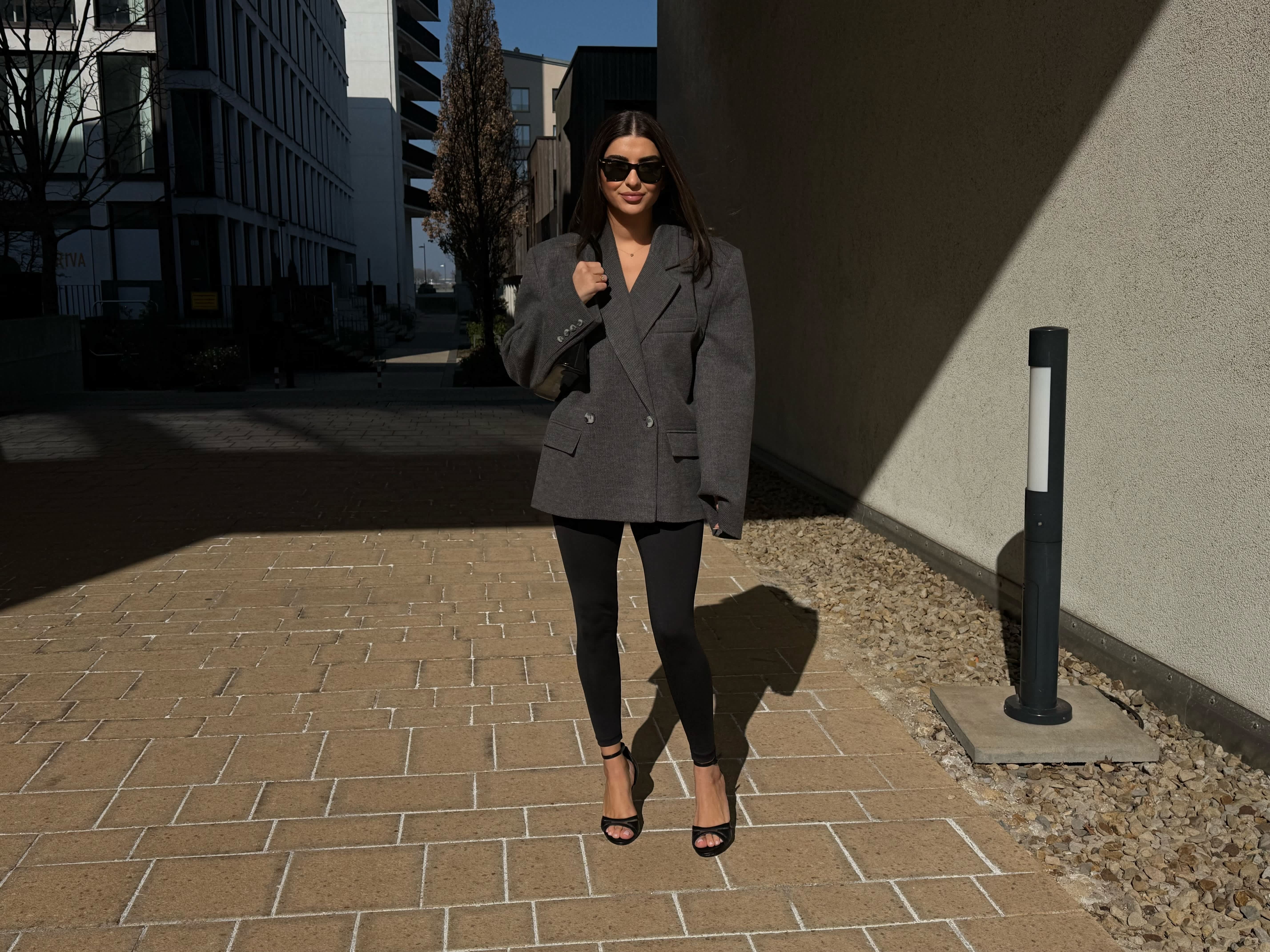 Frau mit schwarzer Leggins, Sandaletten und grauem Blazer mit passender Tasche zwischen zwei Häsuern