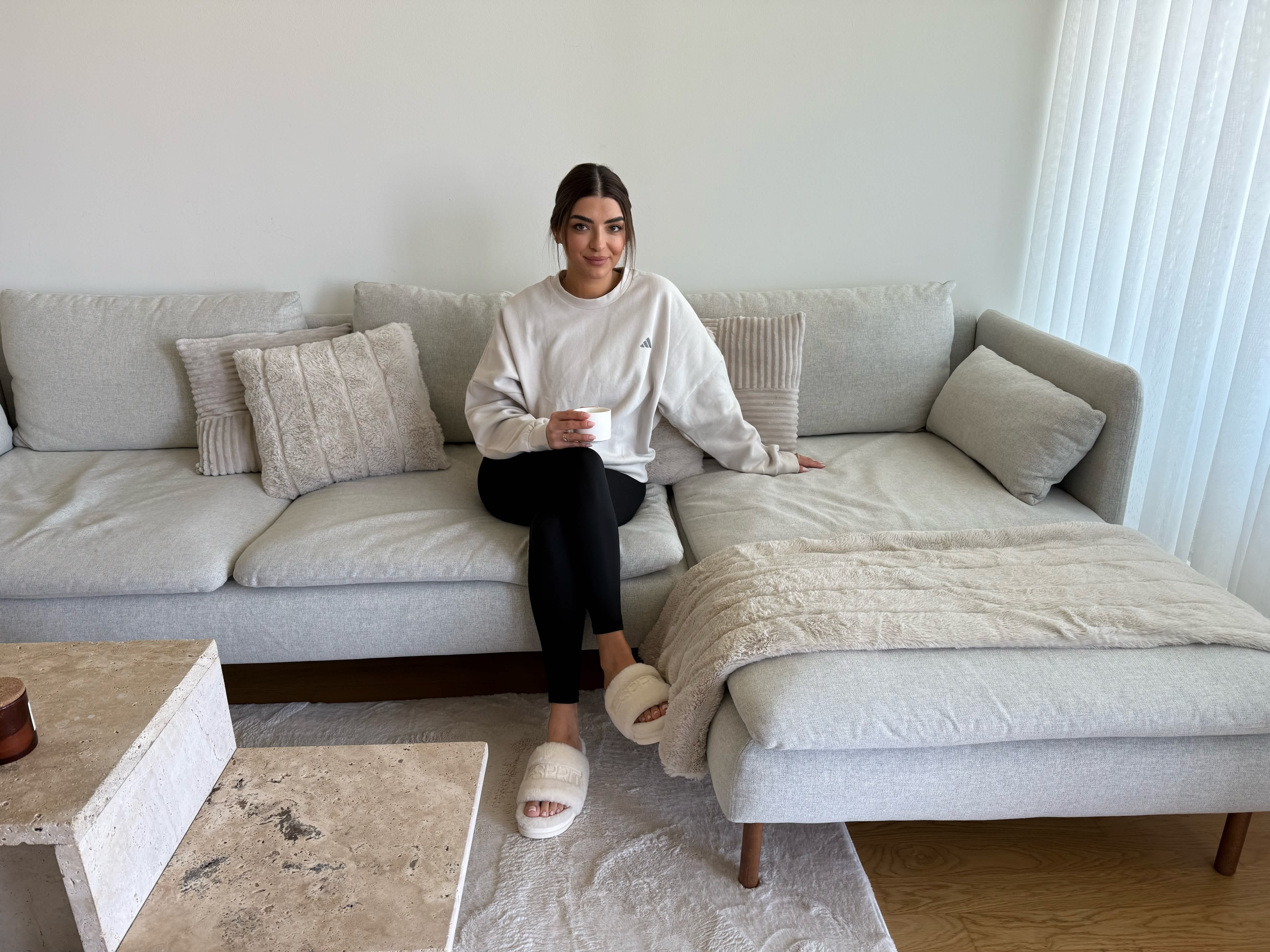 Frau mit schwarzer Leggins, weißen Hausschuhe und hellem Sweater auf hellgrauem Sofa