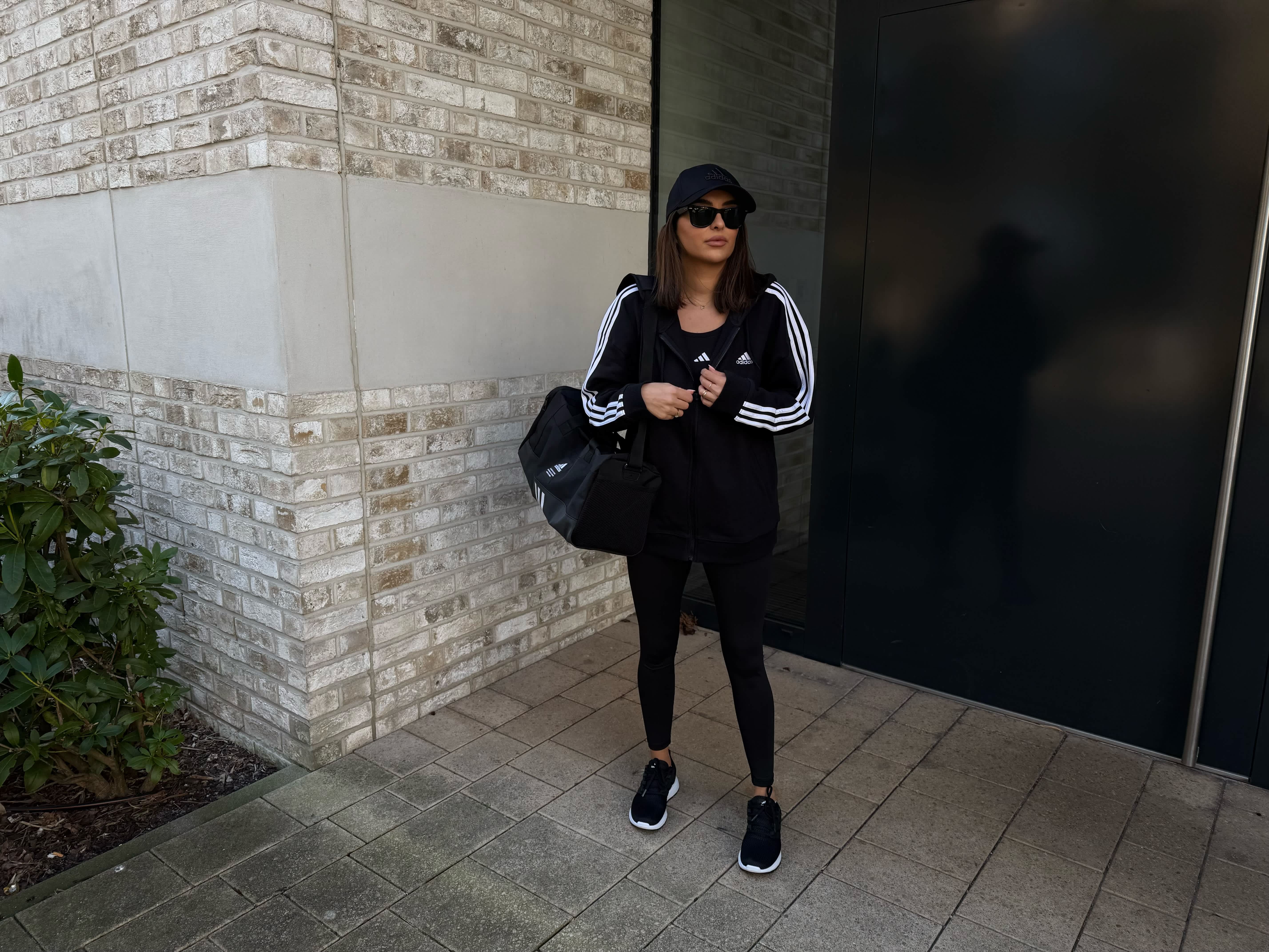 Frau mit schwarzer Leggins, schwarzer Adidas Jacke und passenden Accessoires vor Hauseingang