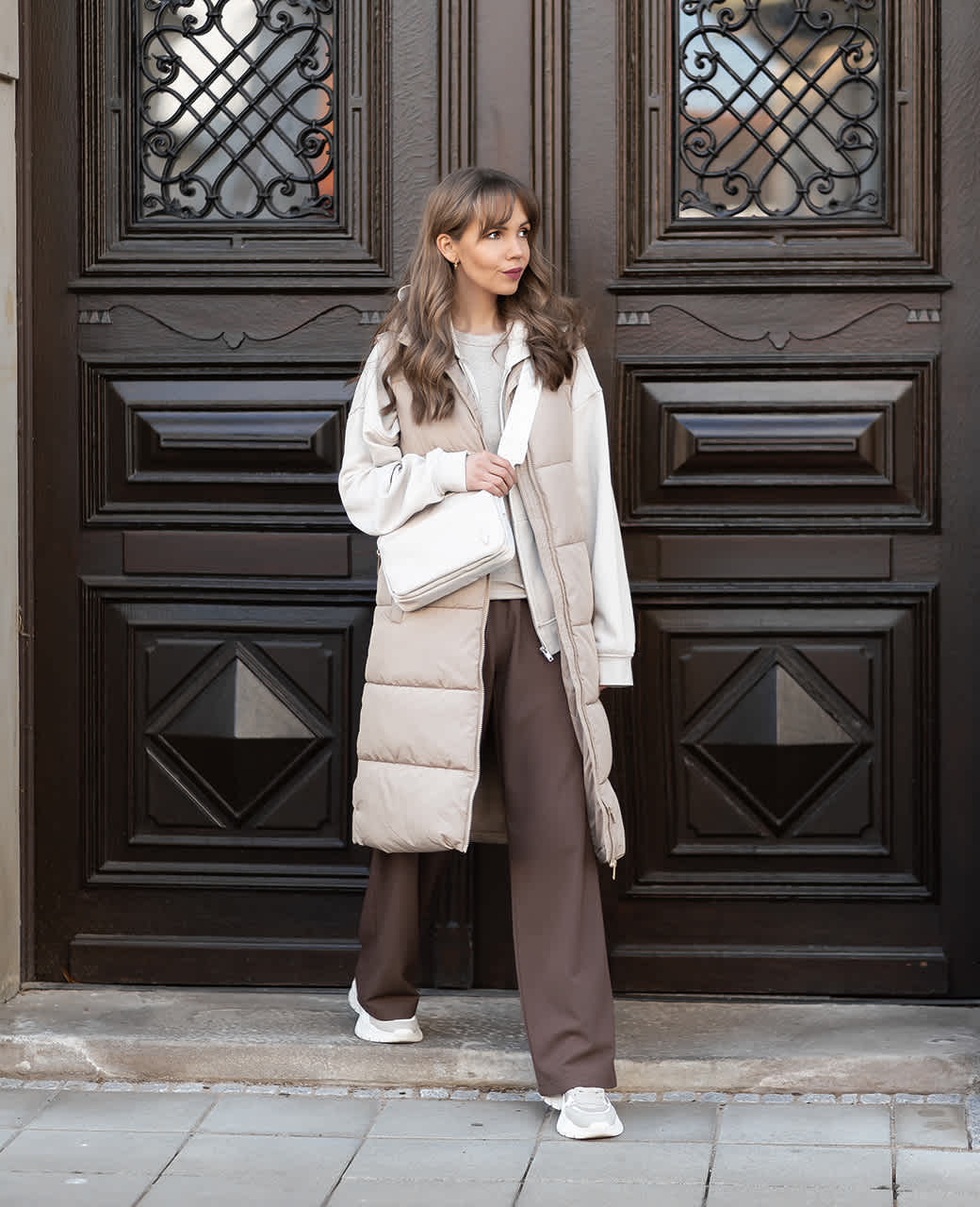 Frau steht vor dunkler Holztür in einer langen, beigefarbenen Steppweste, weitem Sweatshirt, brauner Hose und Chunky-Sneakern. Dazu trägt sie eine weiße Crossbody-Tasche.