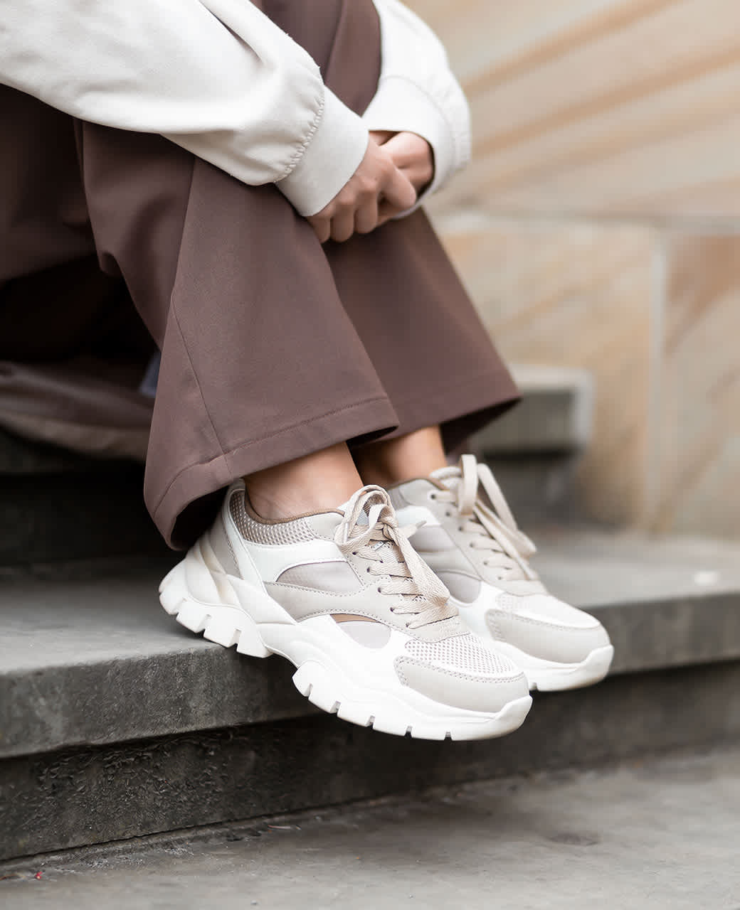 Person sitzt auf Steinstufen in brauner, weiter Hose und trägt beigefarbene Chunky-Sneaker mit heller Sohle.