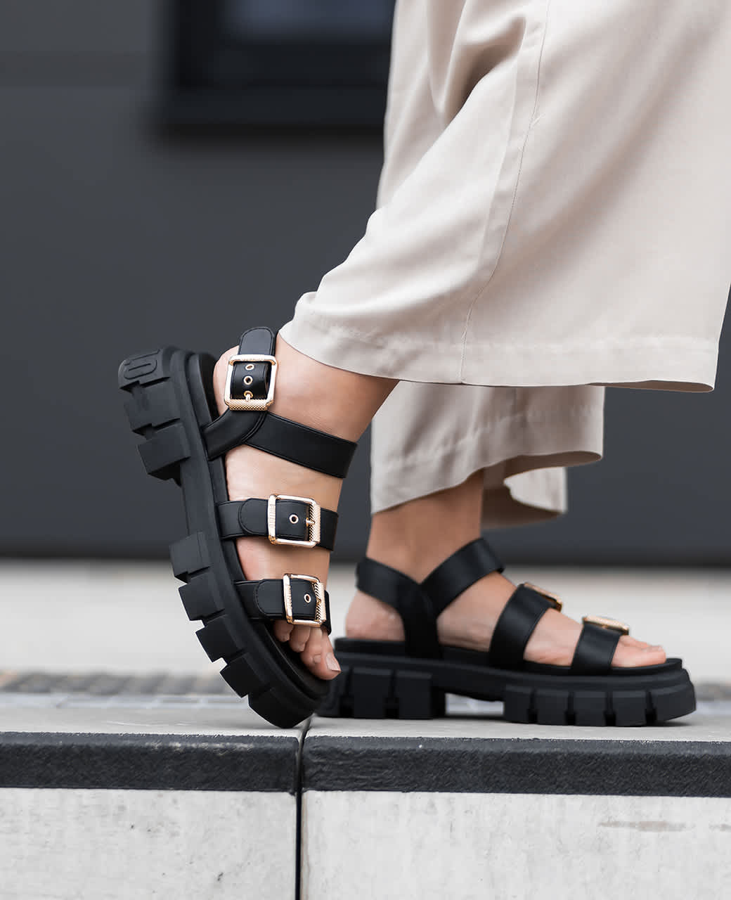 Frau steht auf einem modernen Gehweg in weitem, beigem Hosenstil, schwarzem Blazer und schwarzen Chunky-Sandalen. In der Hand hält sie eine schwarz-weiße Handtasche.