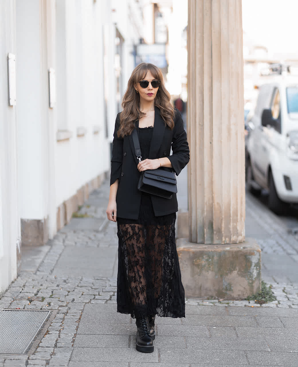 Frau geht auf einem Gehweg entlang, trägt ein schwarzes Spitzenkleid mit Blazer und klobigen Schnürboots. Über der Schulter trägt sie eine schwarze Umhängetasche.