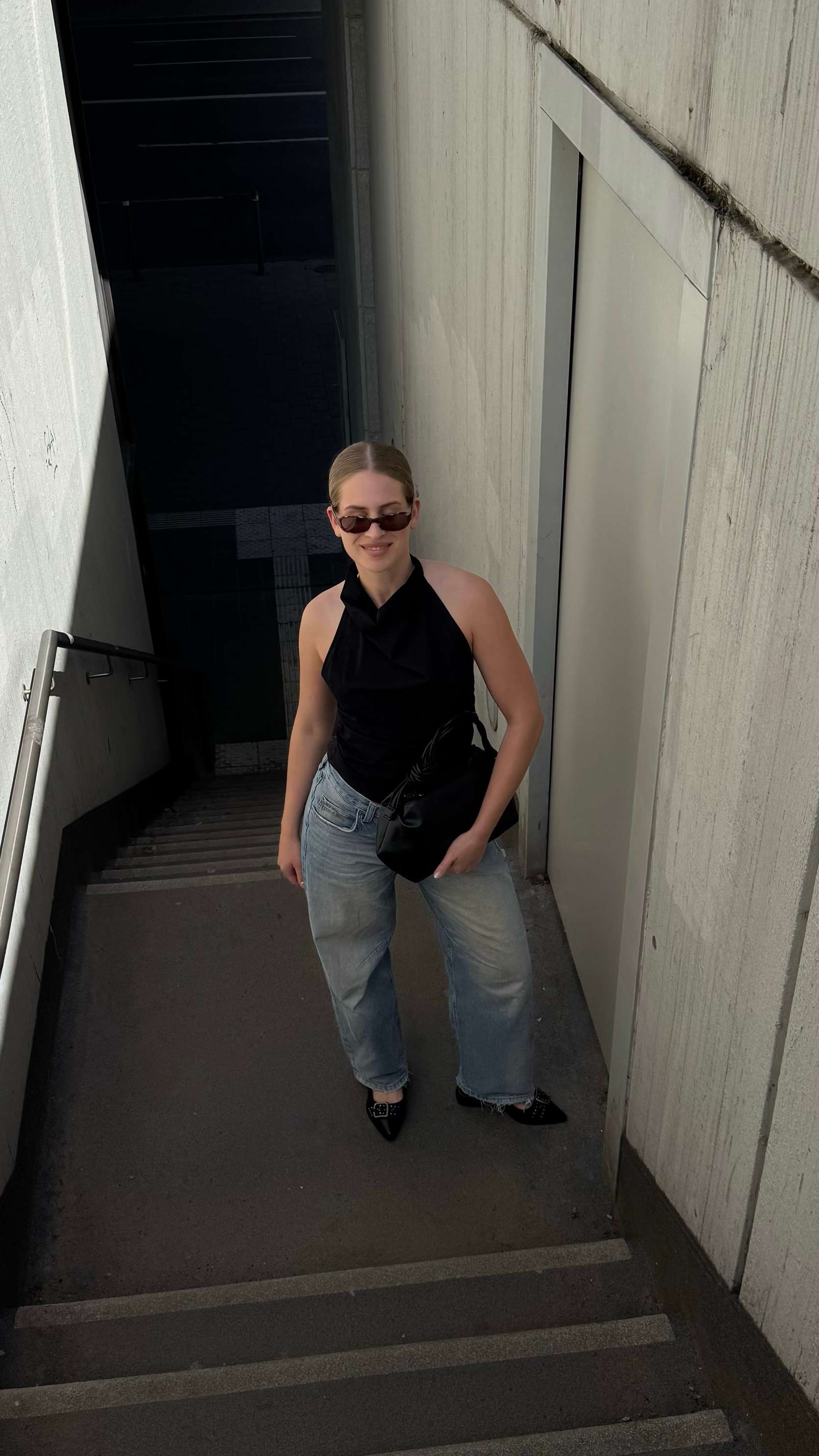 Frau trägt Outfit aus schwarzer Sonnenbrille, schwarzem Off-Shoulder Top mit weiter blauer Jeans und Pointed Flats