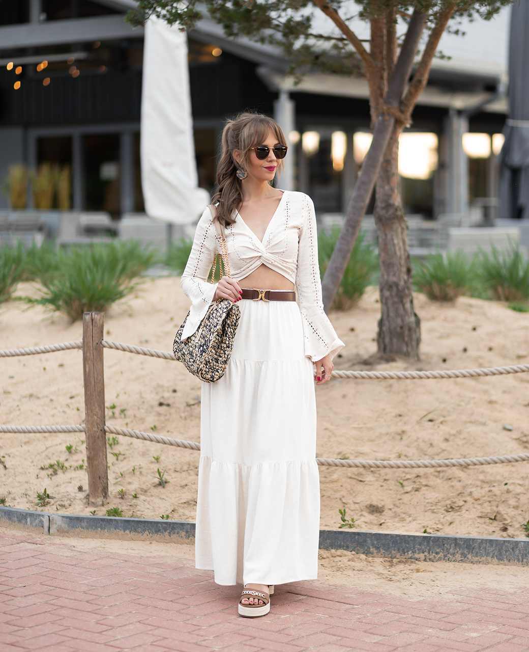Junge Frau in weißem Outfit im Boho-Style, trägt Handtasche und steht vor Strandcafé