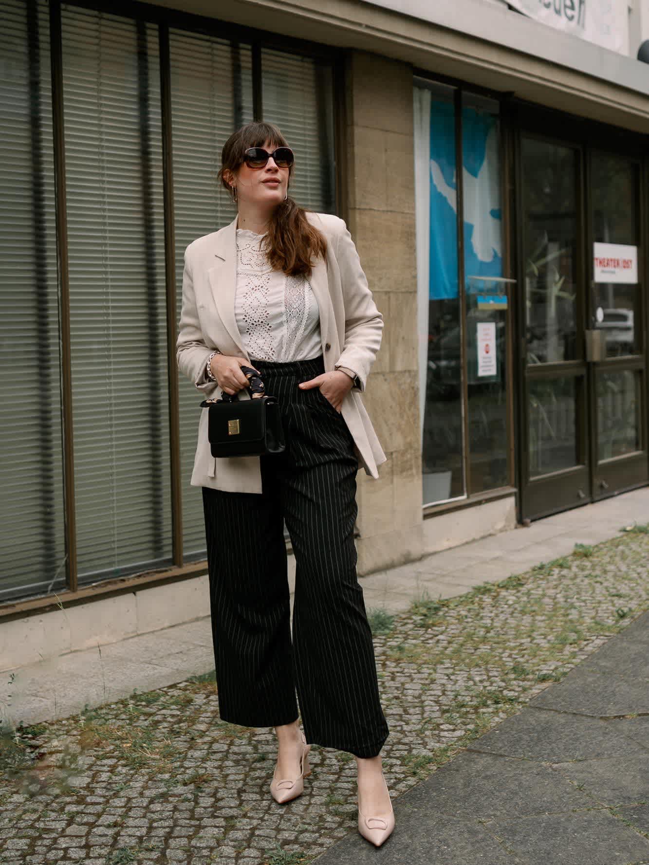 Frau in Office-Outfit, mit schwarzer Sonnenbrille und Handtasche, stehend vor Gebäude