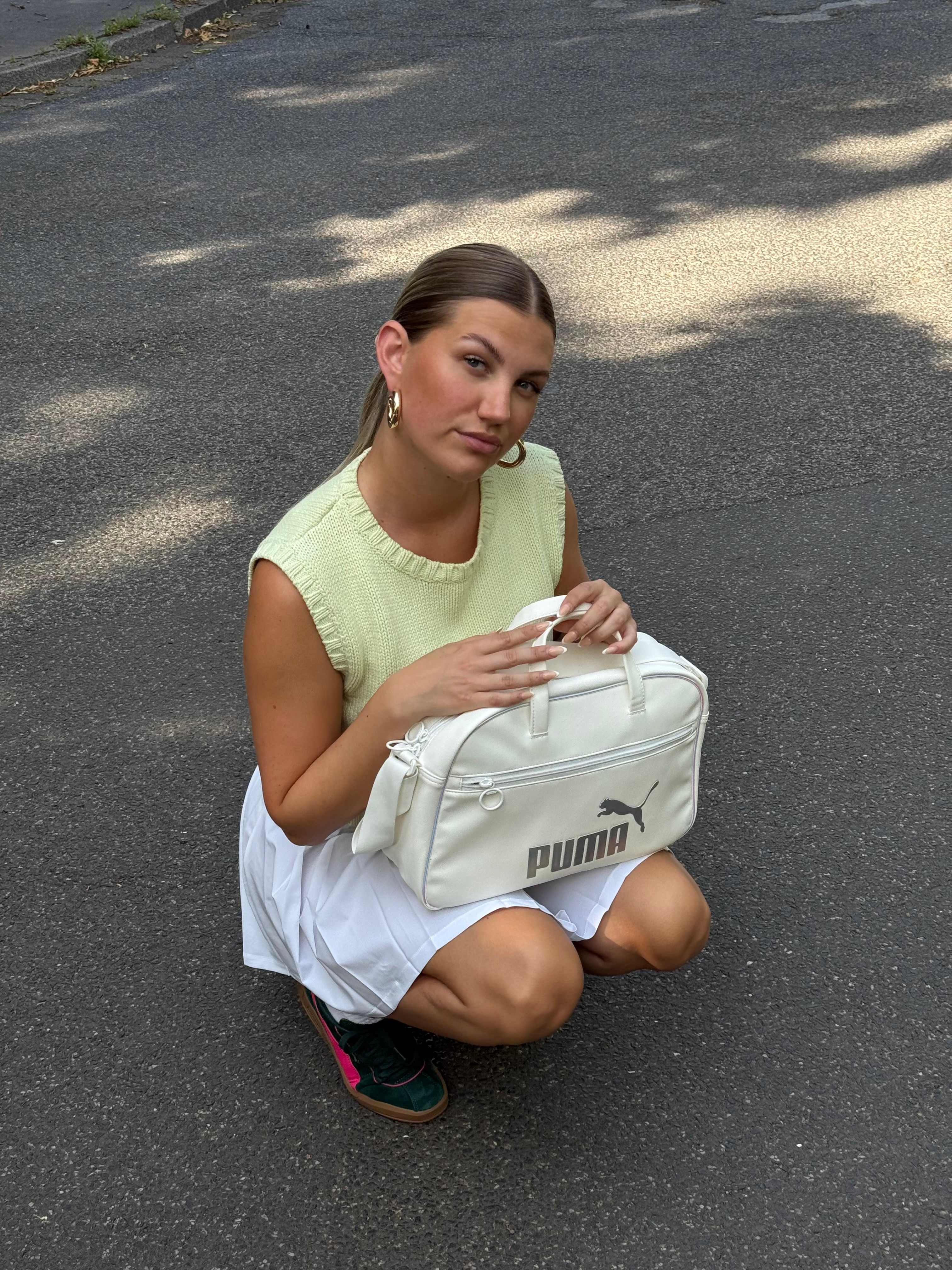 Frau mit gelbem Shirt und weißem Tennis-Outfit, mit Puma, Tasche, hockt auf Asphalt